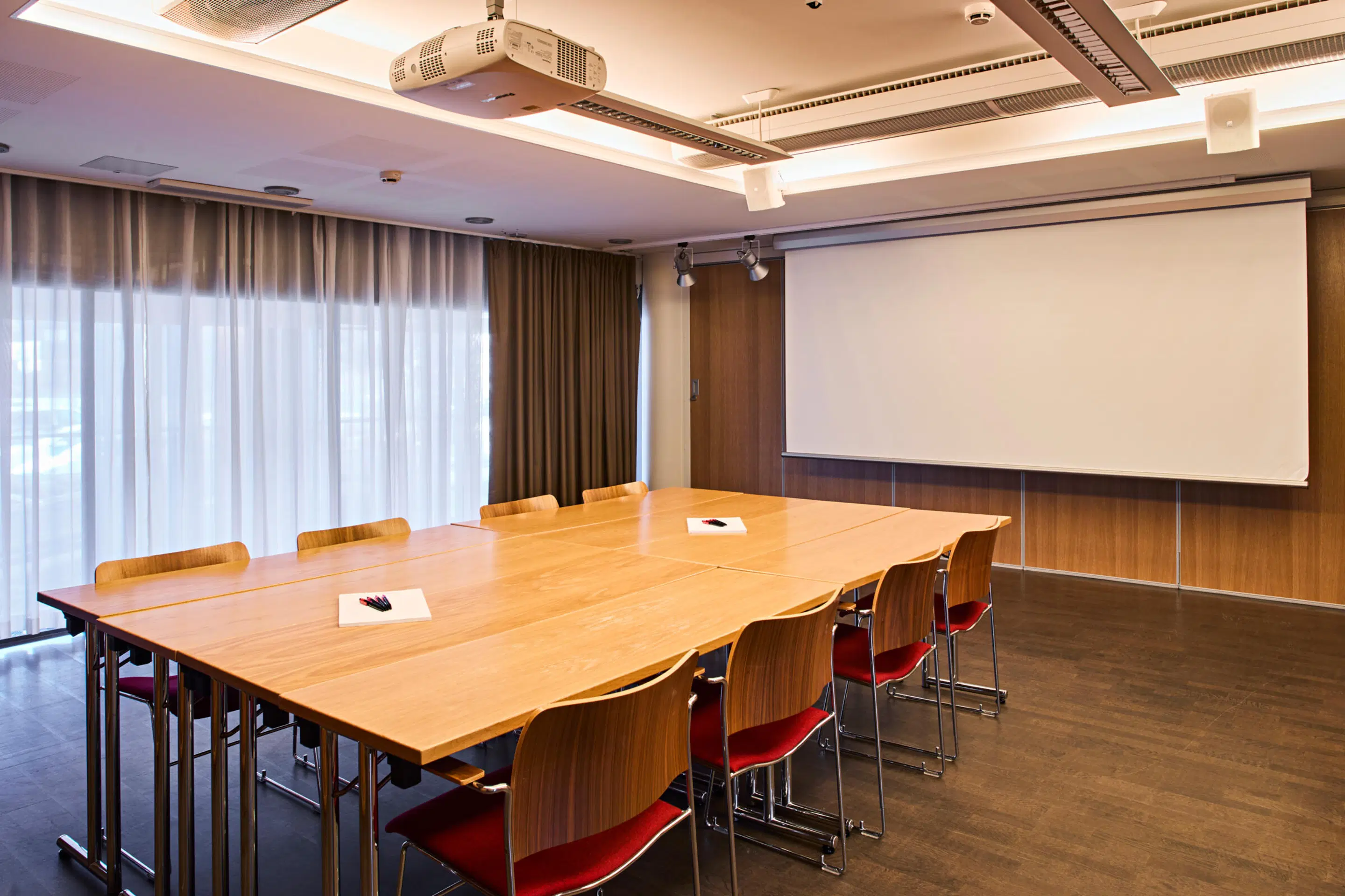 Conference room with wooden table and chairs, ceiling projector and projection screen on wall. Room has level flooring and large curtains.