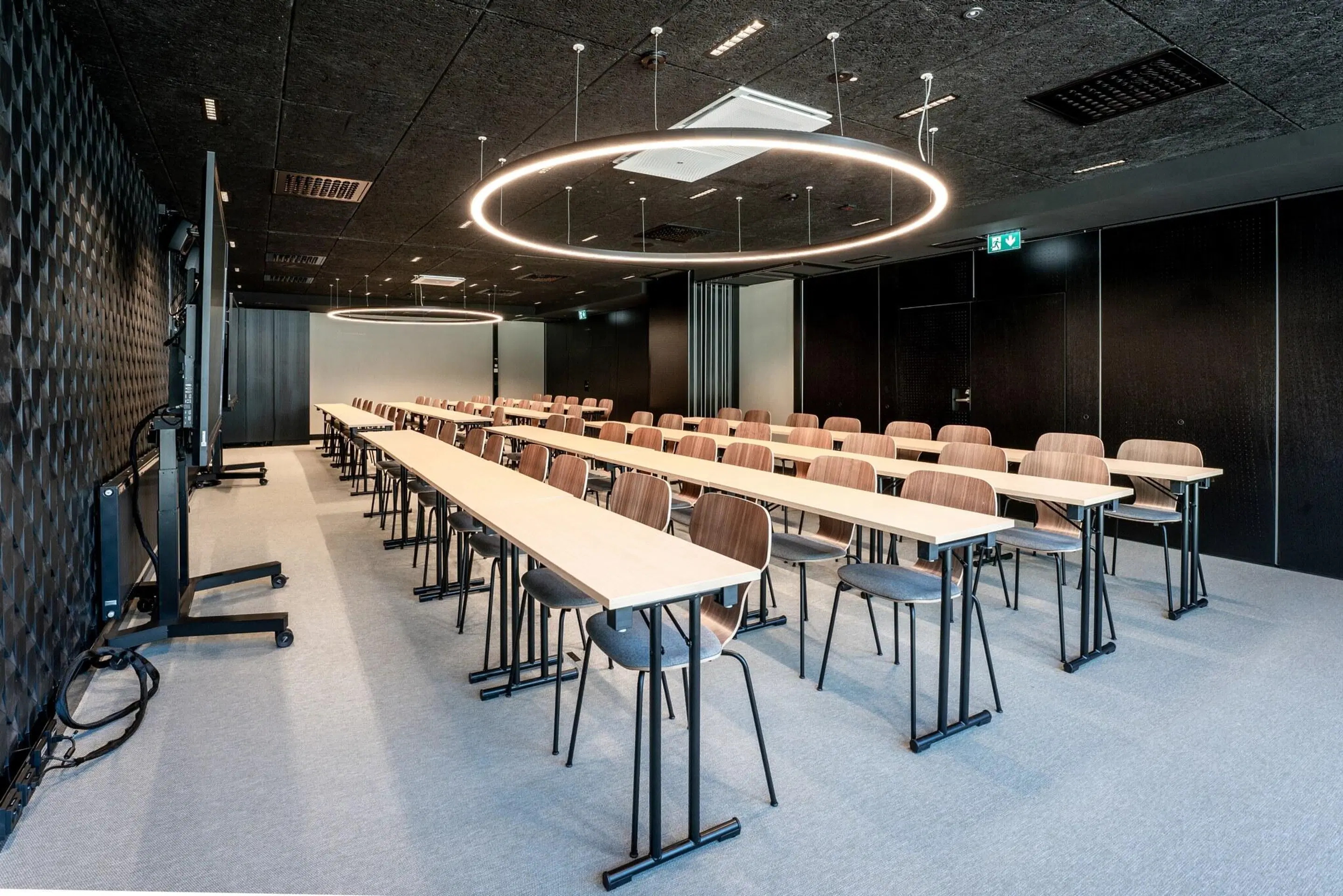 Modern conference room with long tables and wooden chairs, circular ring lights on ceiling, level floor surface and dark walls.