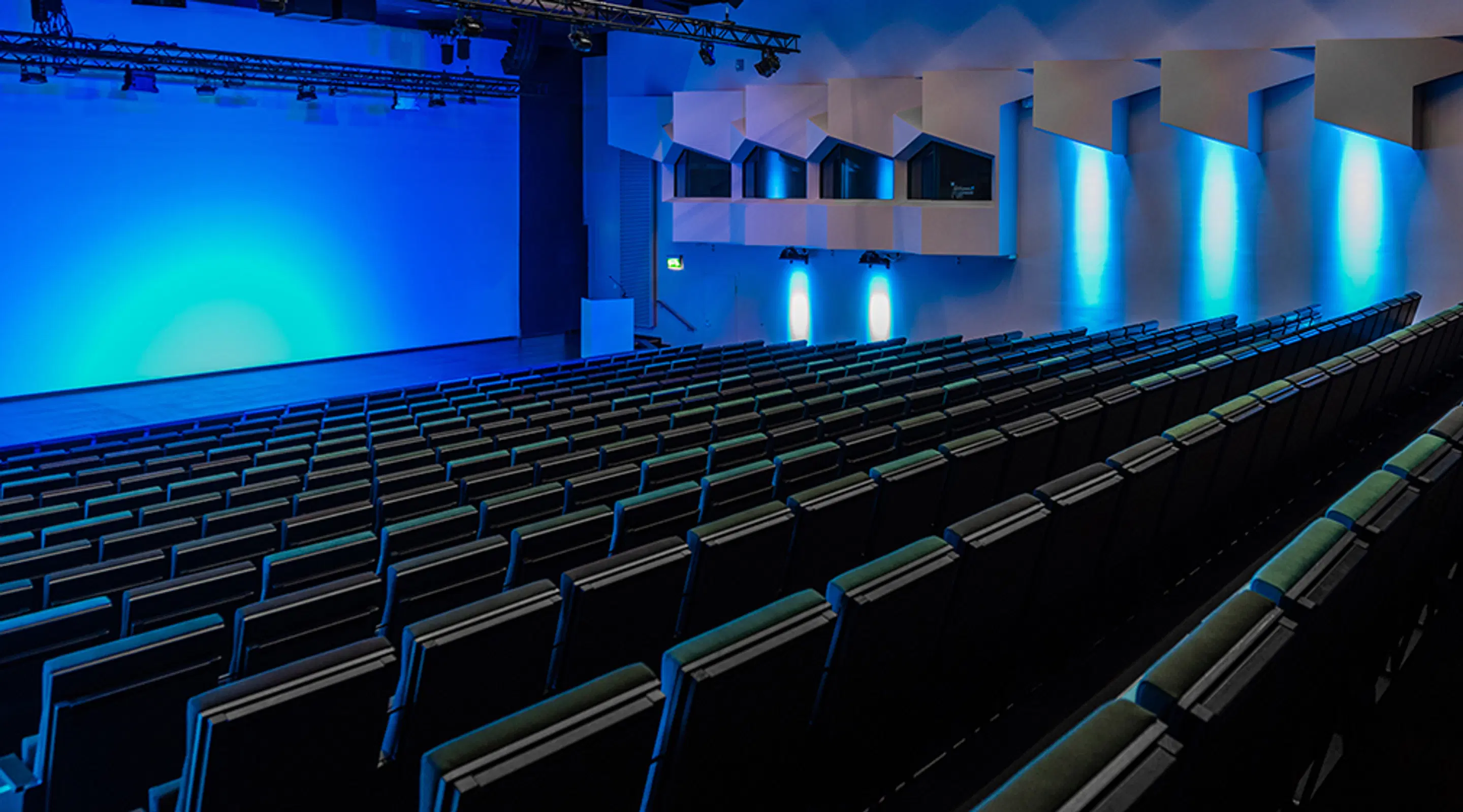 Auditorium med sluttande stolarader, bred mittgång och en scen upplyst i blått ljus.