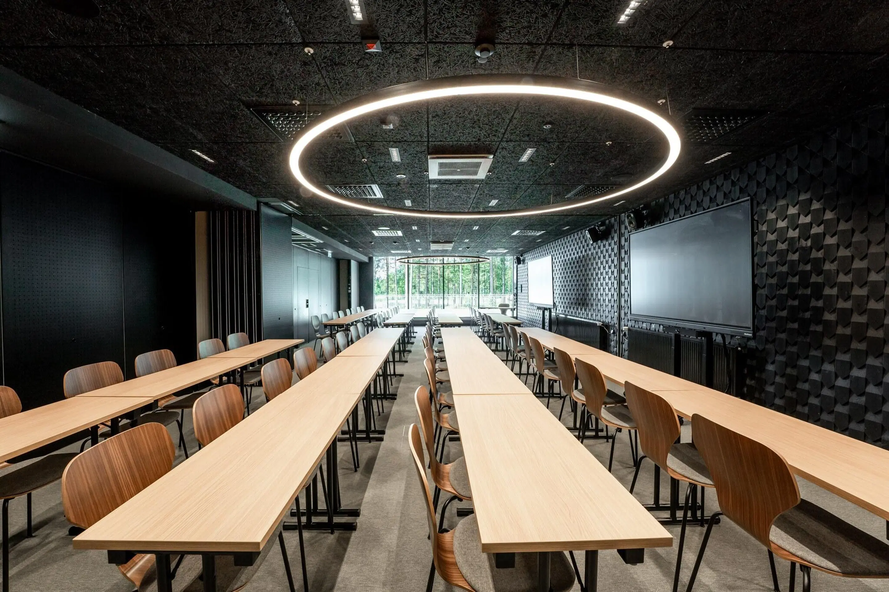 Modern conference room with wooden tables in rows, circular light ring on ceiling, screens on walls, and large windows with nature view.