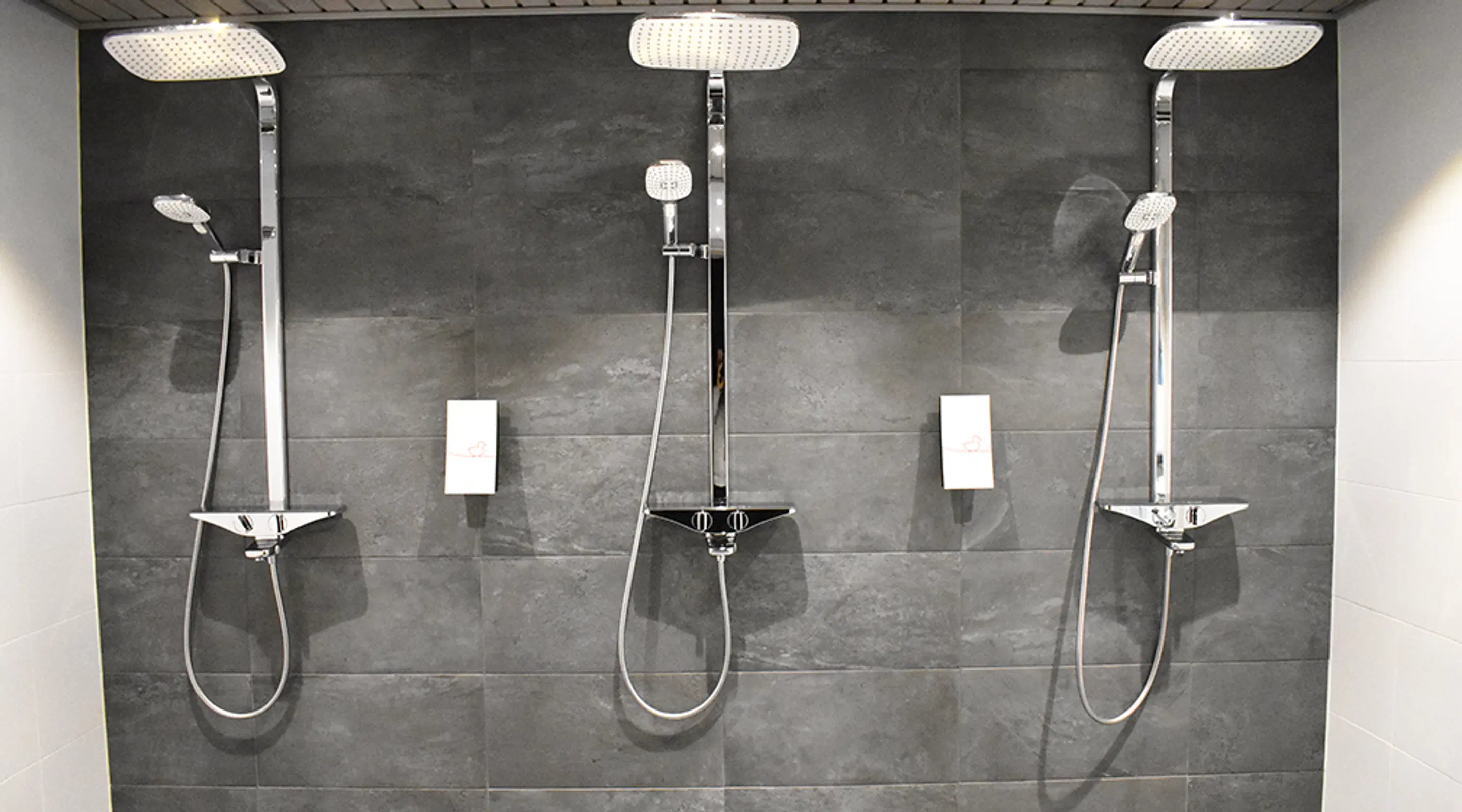 Three modern showers in a row on a grey tiled bathroom wall, each with shiny shower heads and handheld shower attachment.