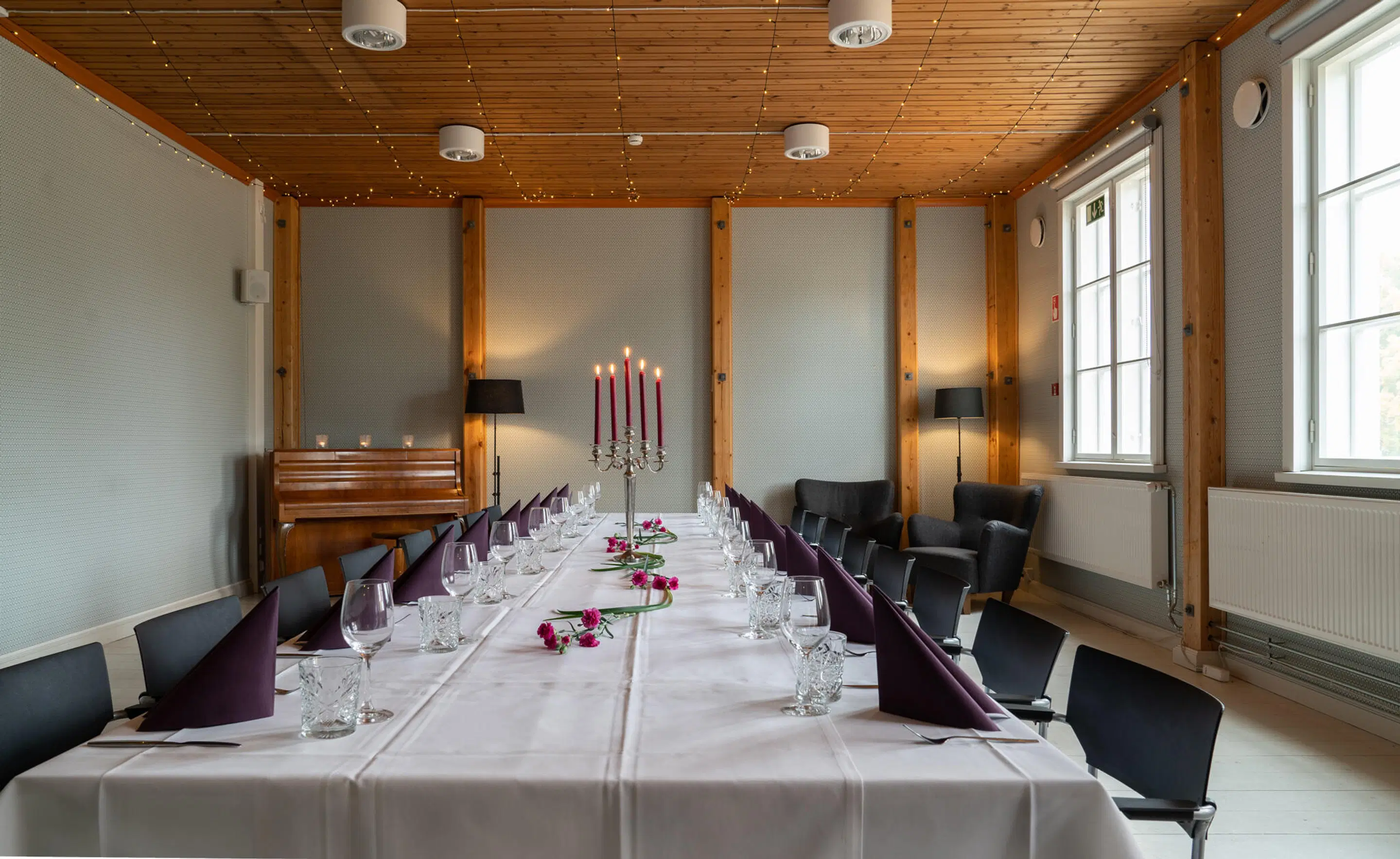 Long table formally set with candelabra and wine glasses. Room features wooden ceiling with string lights, large windows, and a piano.