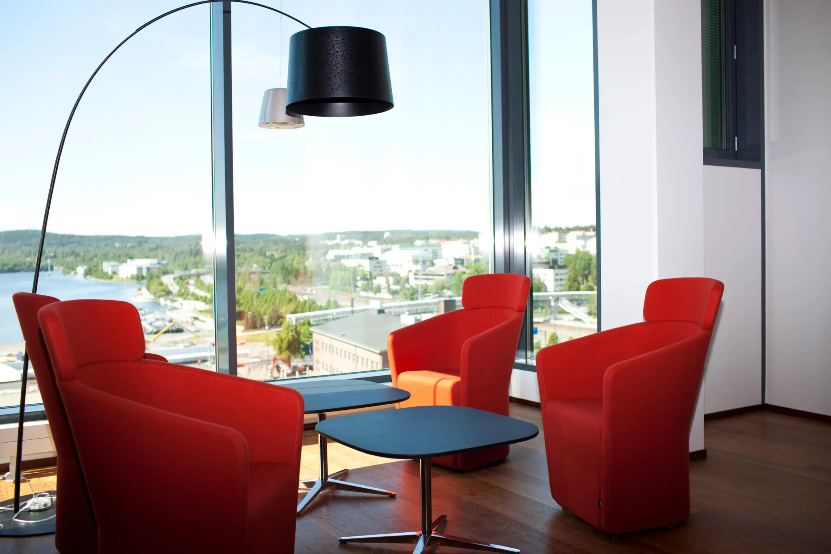Lounge area with red armchairs, blue coffee table and arc floor lamp. Wooden flooring and large windows overlooking city and water views.