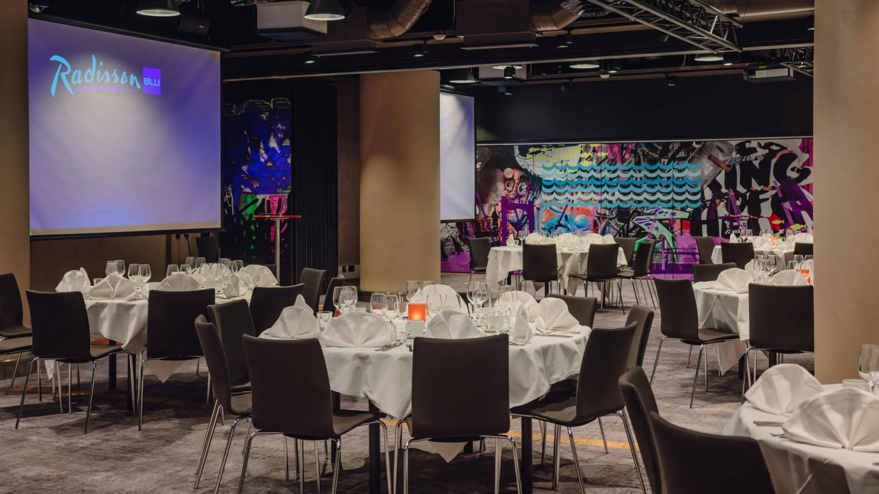 Radisson Blu hotel banquet room with round tables covered in white tablecloths, two projection screens and graffiti artwork on wall. Level floor and open space.