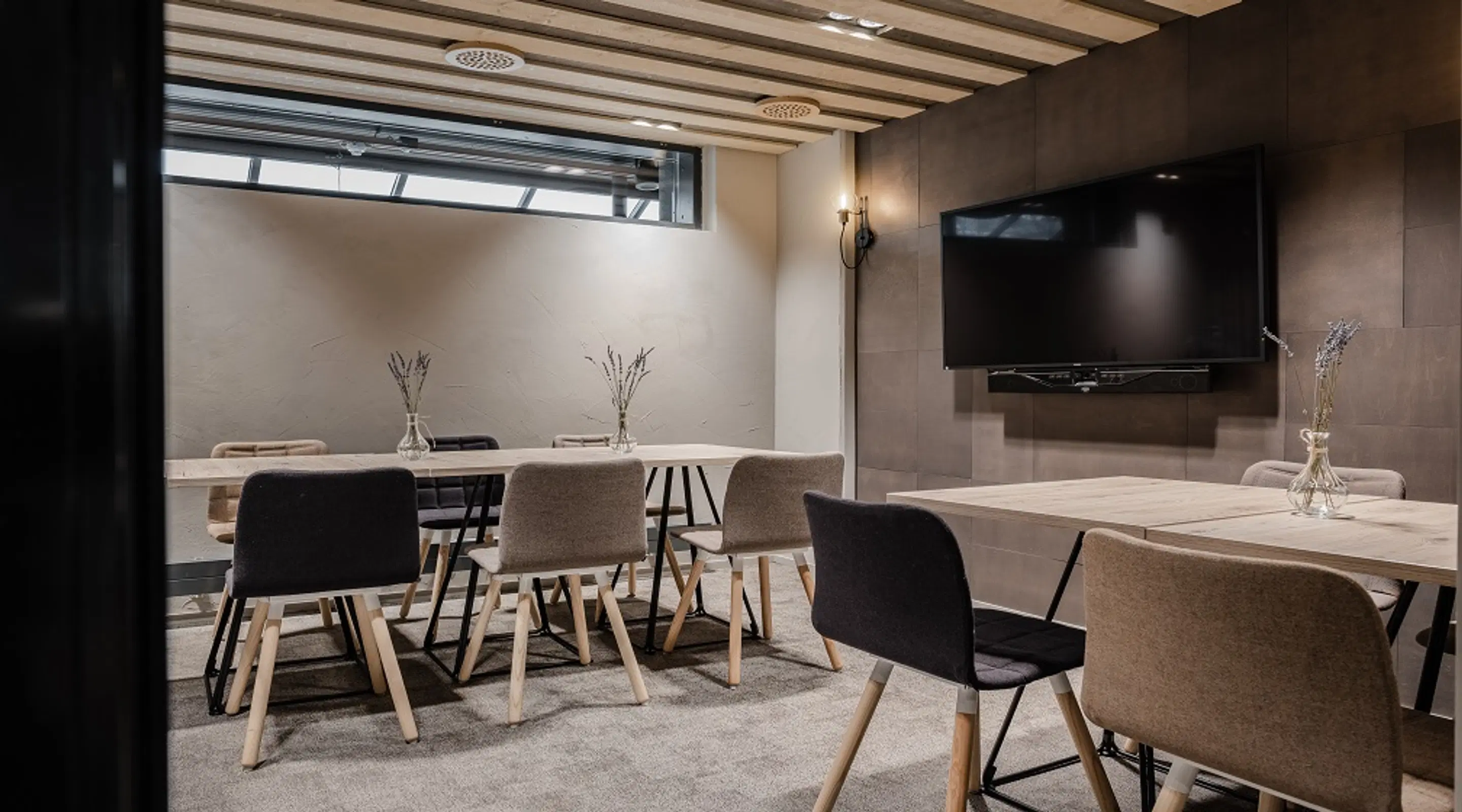 Moderni kokoushuone, jossa kaksi puupöytää, tuoleja, seinään asennettu televisio ja ikkunat ylhäällä. Lattia on pehmeä matto.