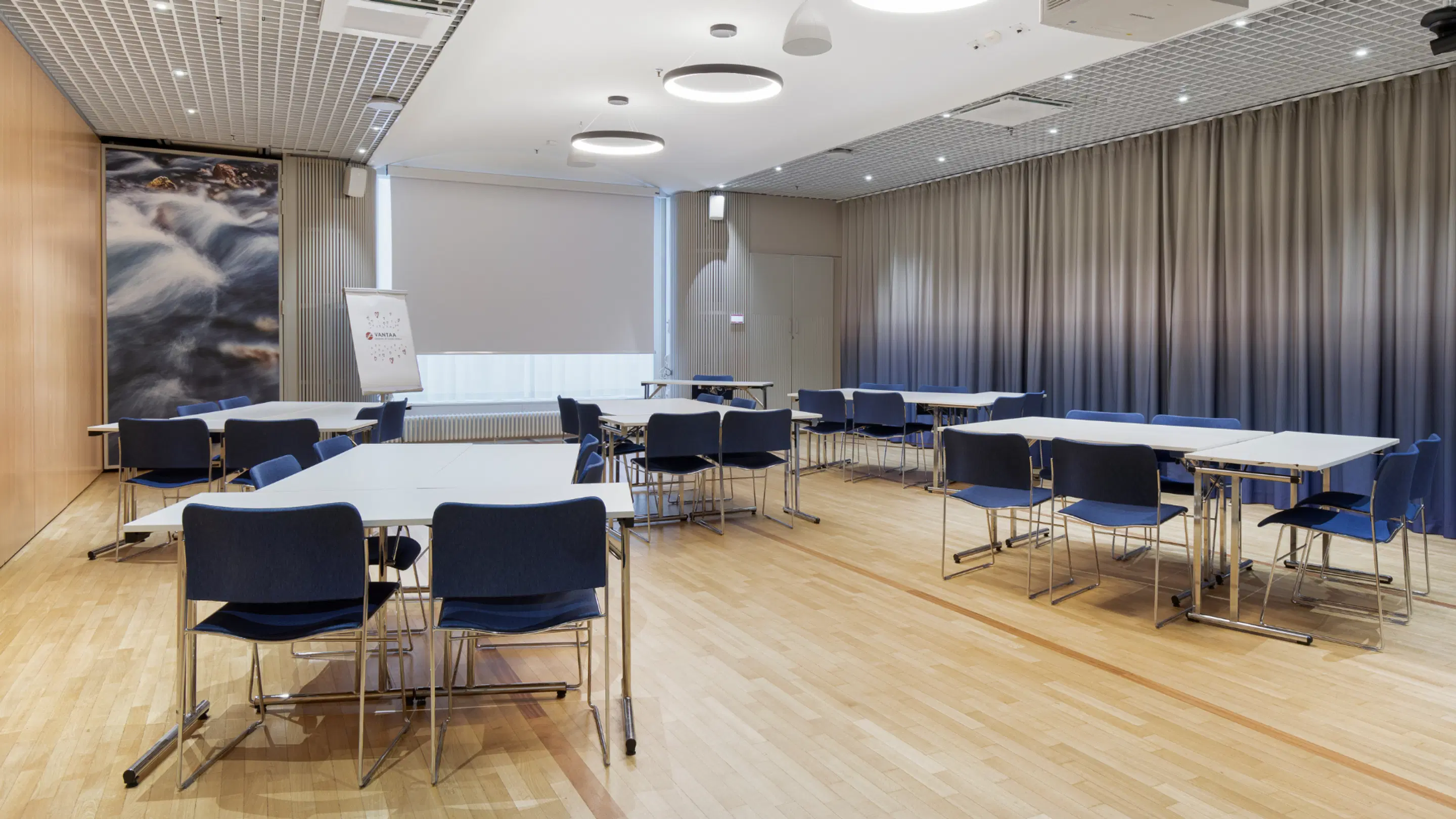 Modern meeting room with wooden floor and blue chairs. Bright space with grouped tables, whiteboard at front and nature image on wall.