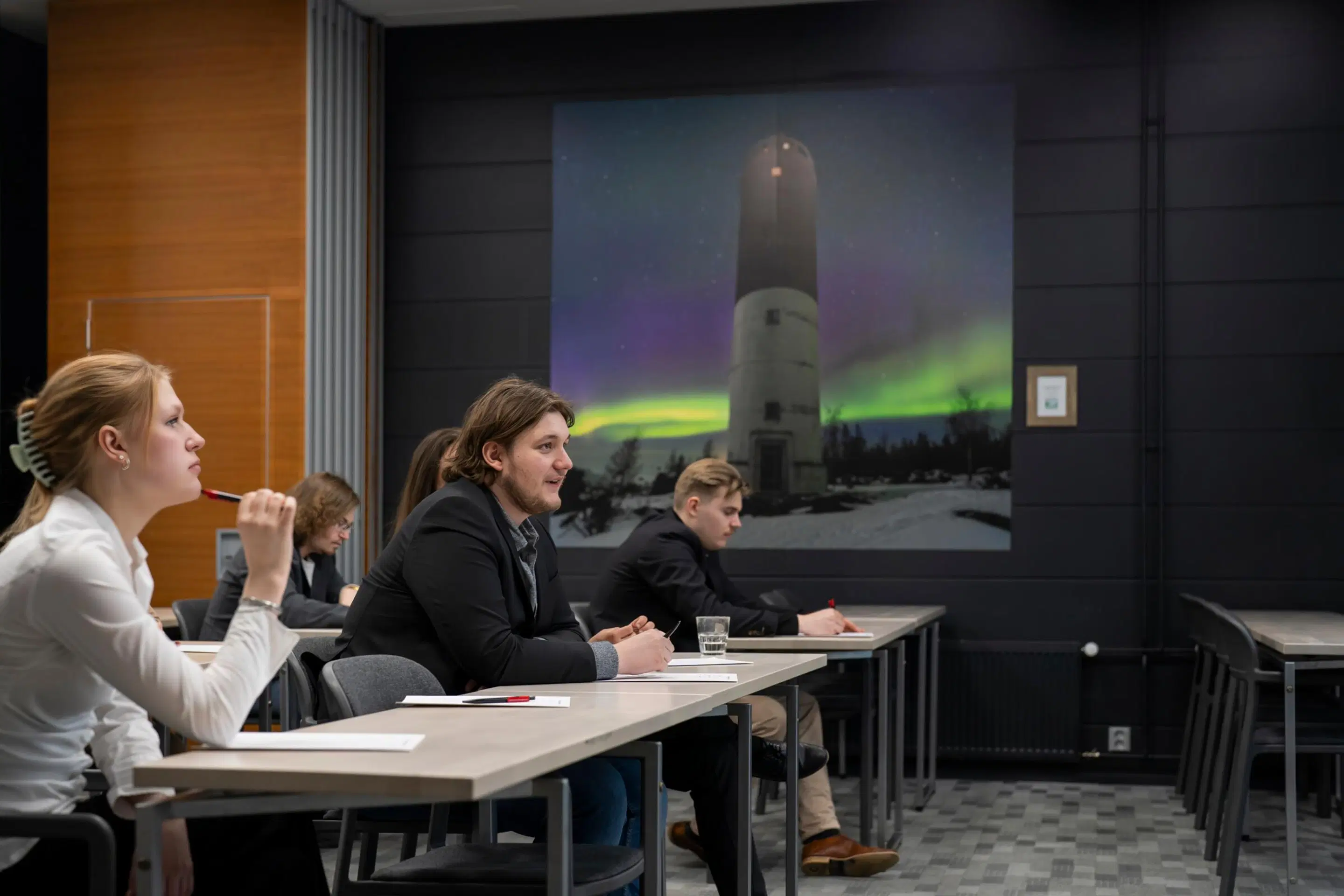 Students sitting at tables in a lecture room. A large image of a lighthouse under northern lights is displayed on the wall.