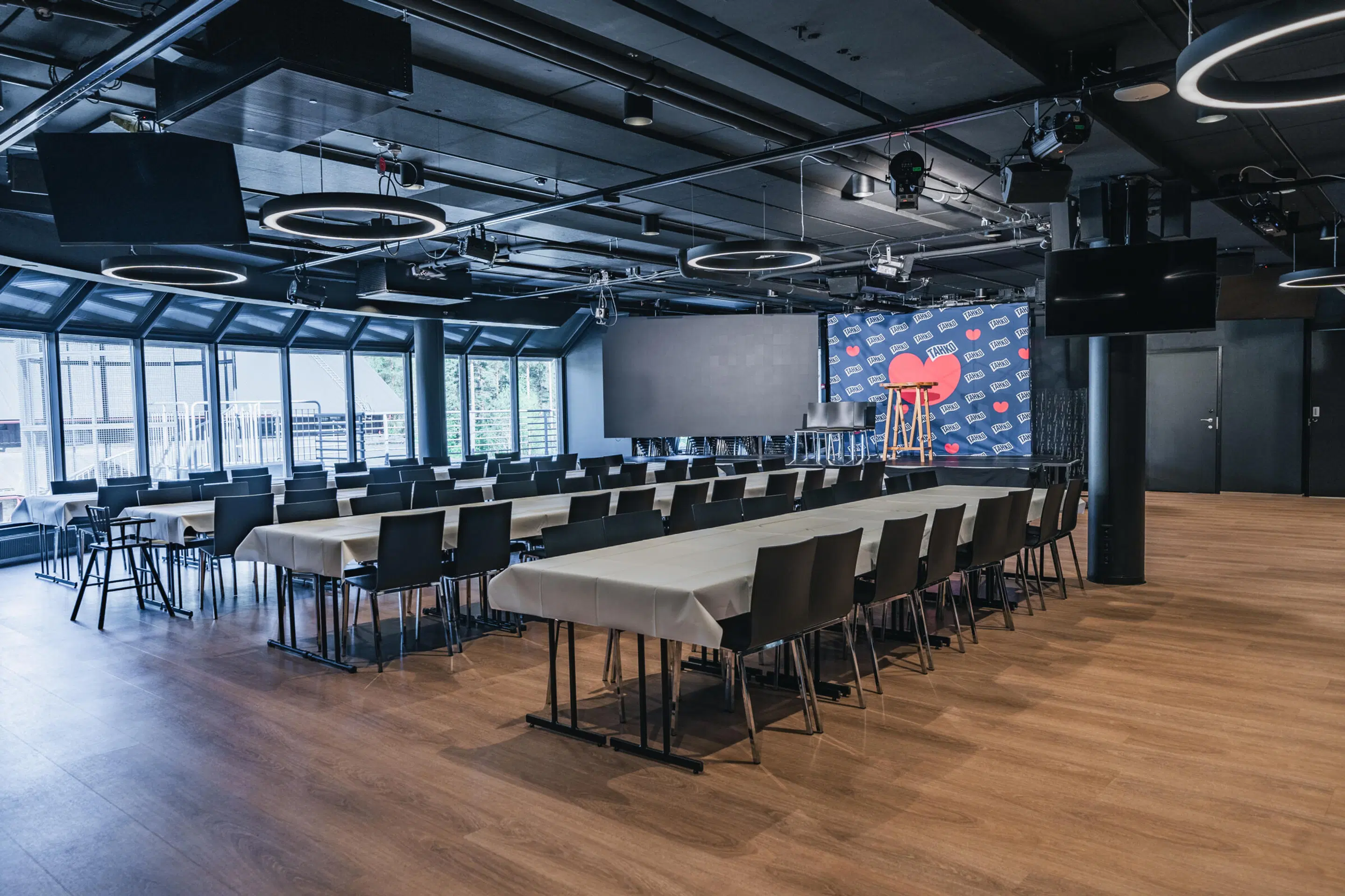 Moderni konferenssihuone, jossa pöydät tuoleineen, lavan edessä näyttö ja taustaseinä. Puulattia, isot ikkunat ja rengasmaiset valaisimet katossa.