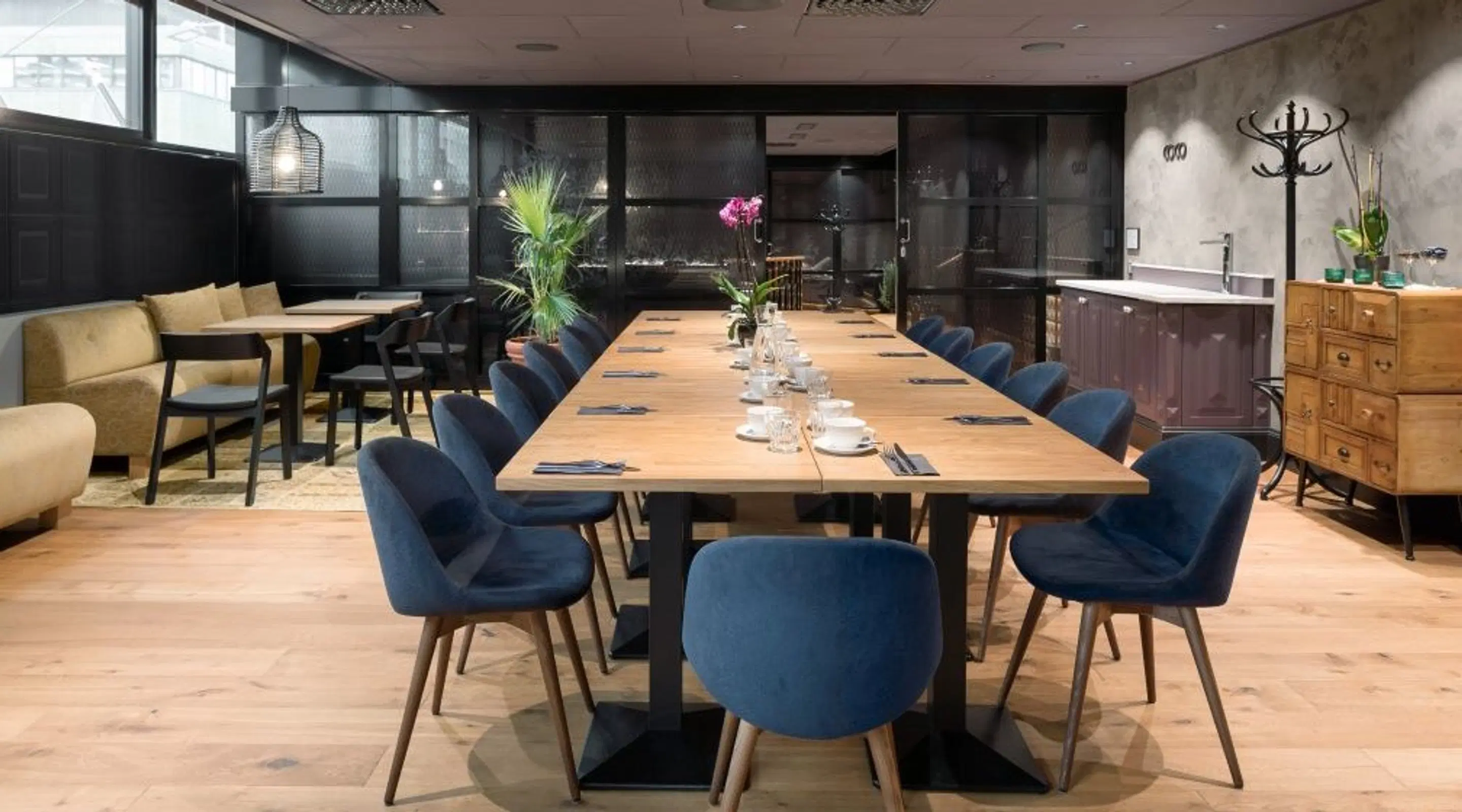Stylish meeting room with long wooden table surrounded by blue chairs, small kitchen area and seating nook. Space features wooden flooring and plants.