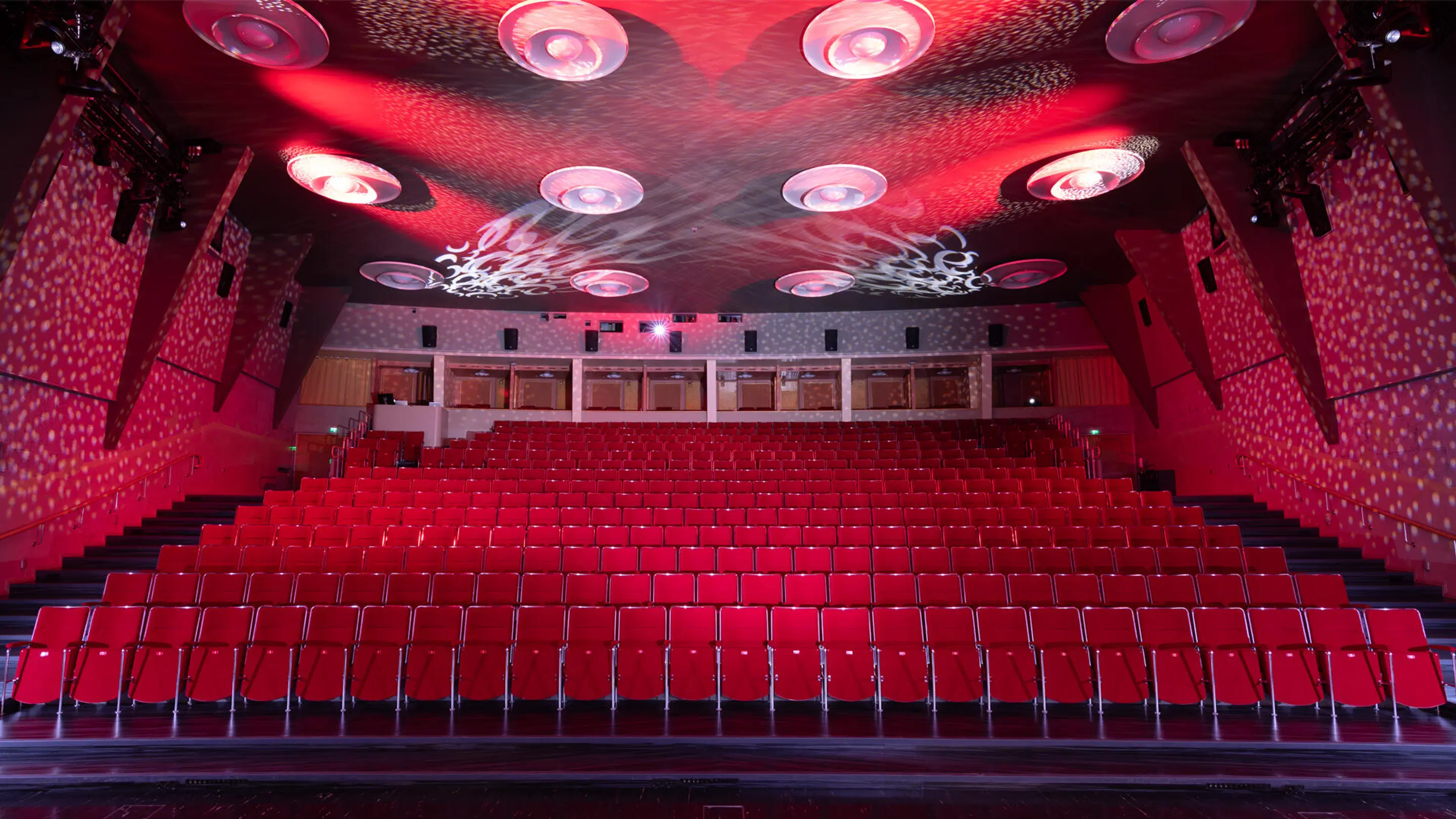 Empty auditorium with bright red seats, stepped aisles, and decorative ceiling lights casting red patterns.