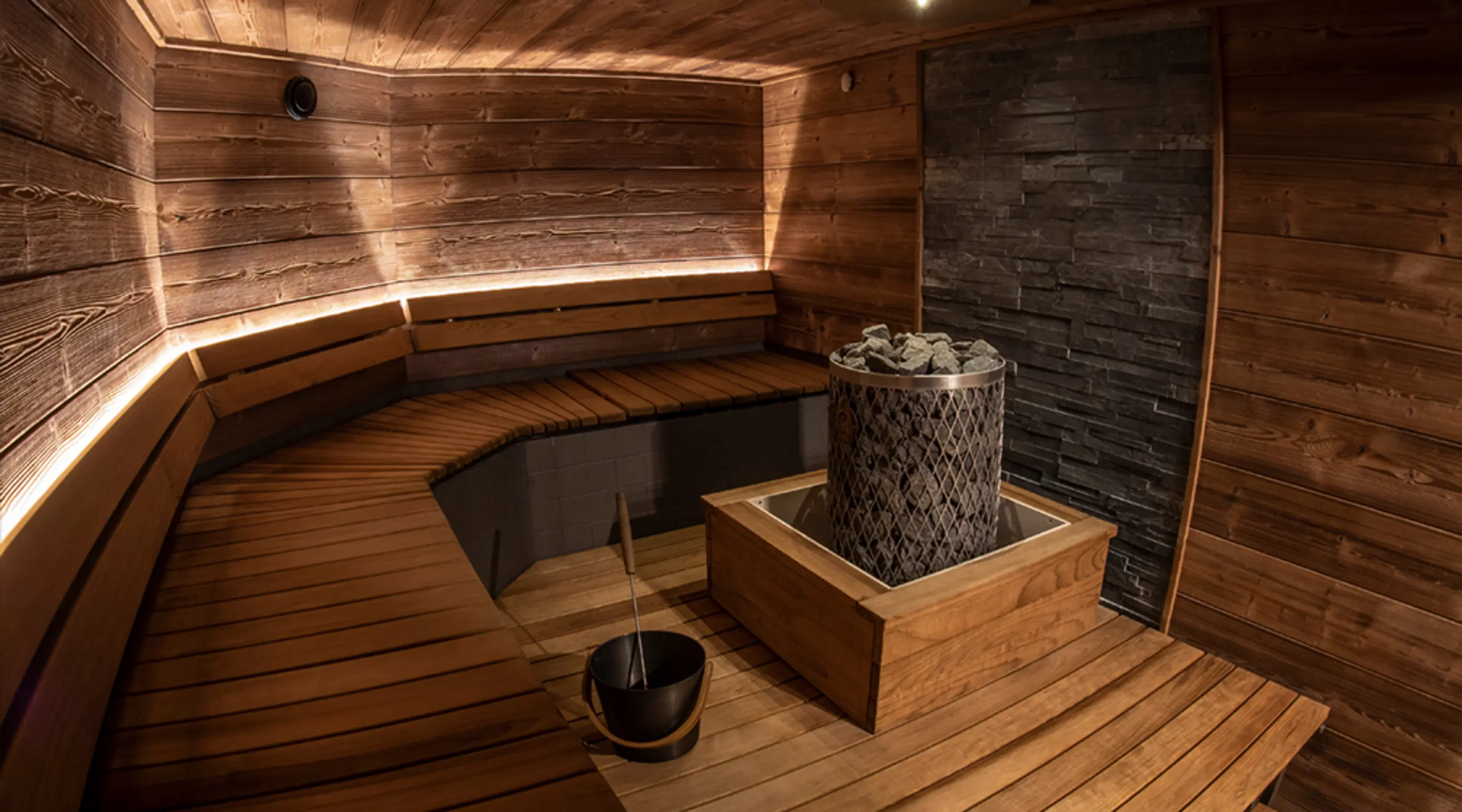 Modern sauna with wooden interior, stone tiles around the stove. Walls feature indirect lighting and L-shaped benches.