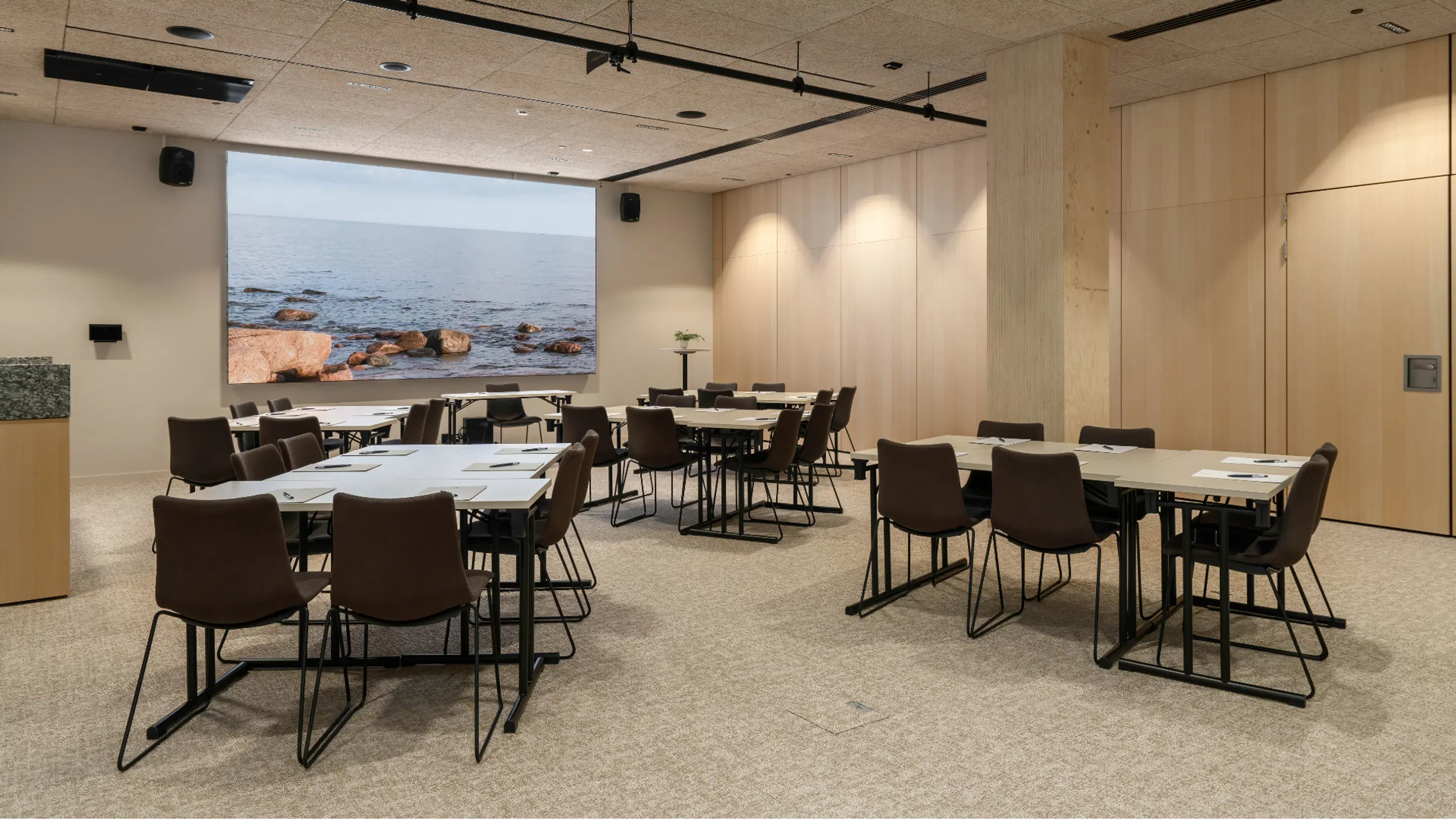 Modern meeting room with light wooden panel walls, table arrangements and large sea-themed projector screen. Level carpeted floor and good lighting.