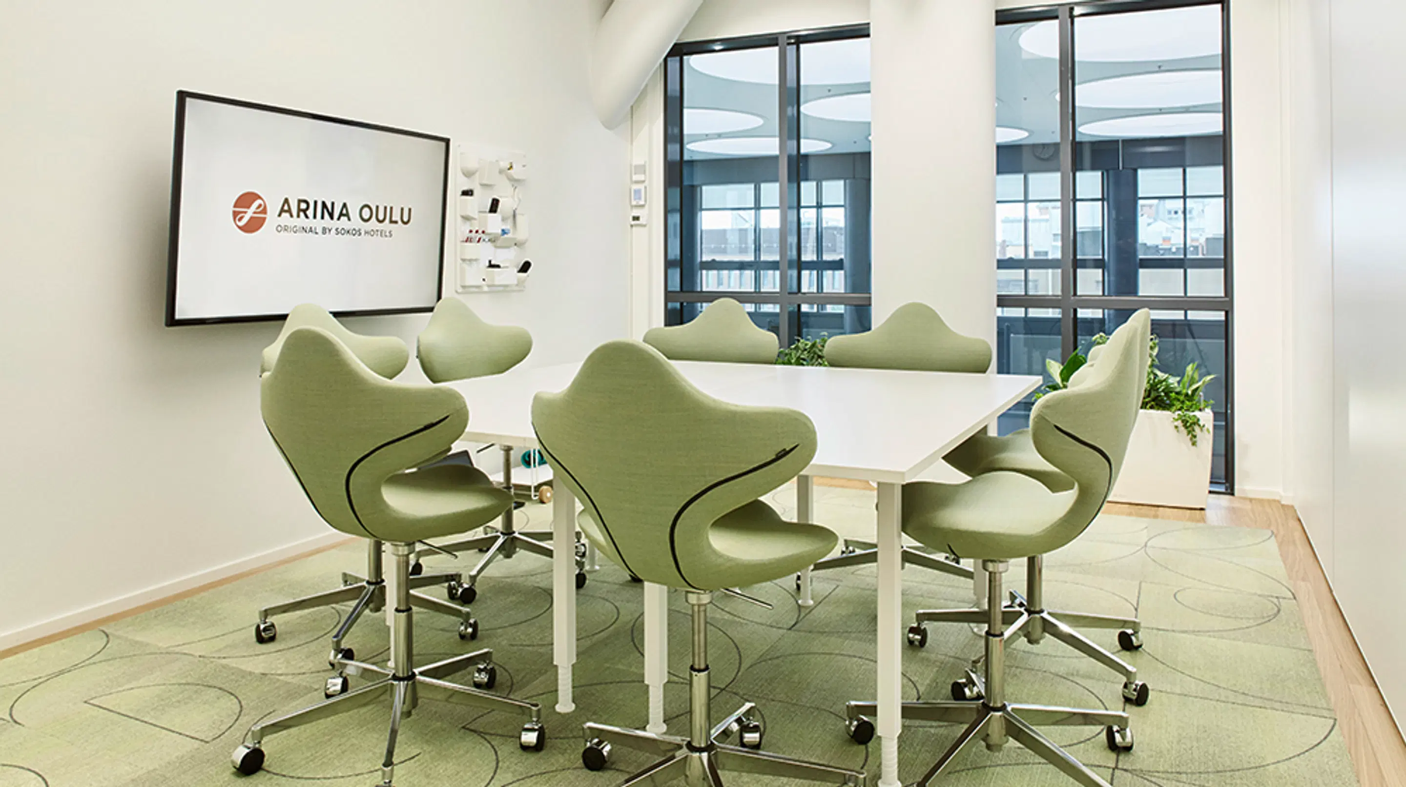 Modern meeting room with green ergonomic chairs around a white table. Bright interior with green carpet and a display screen on the wall.