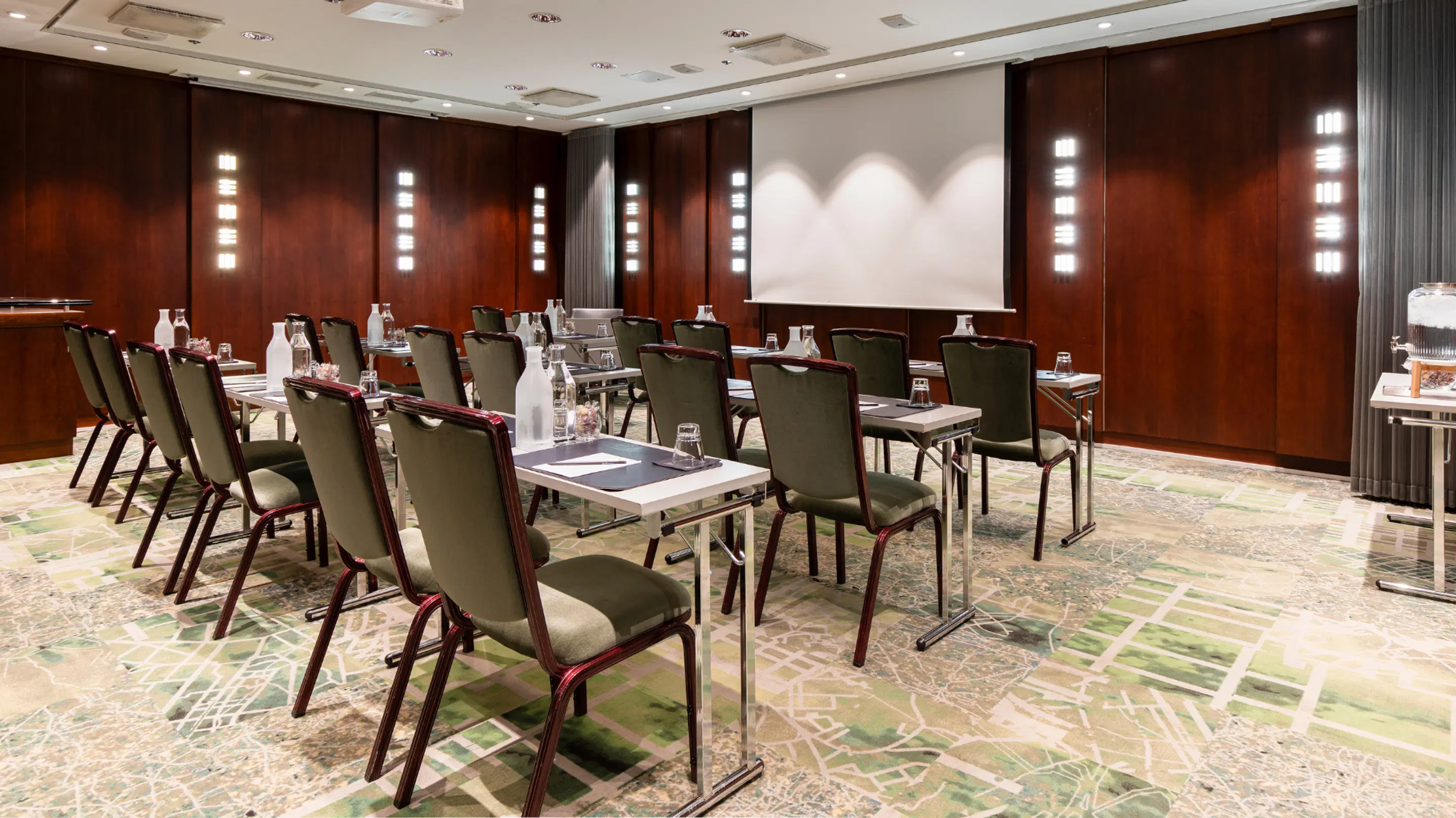 Conference room with dark wood paneling, green chairs and white tables arranged in rows. Projection screen at front. Patterned, flat floor.