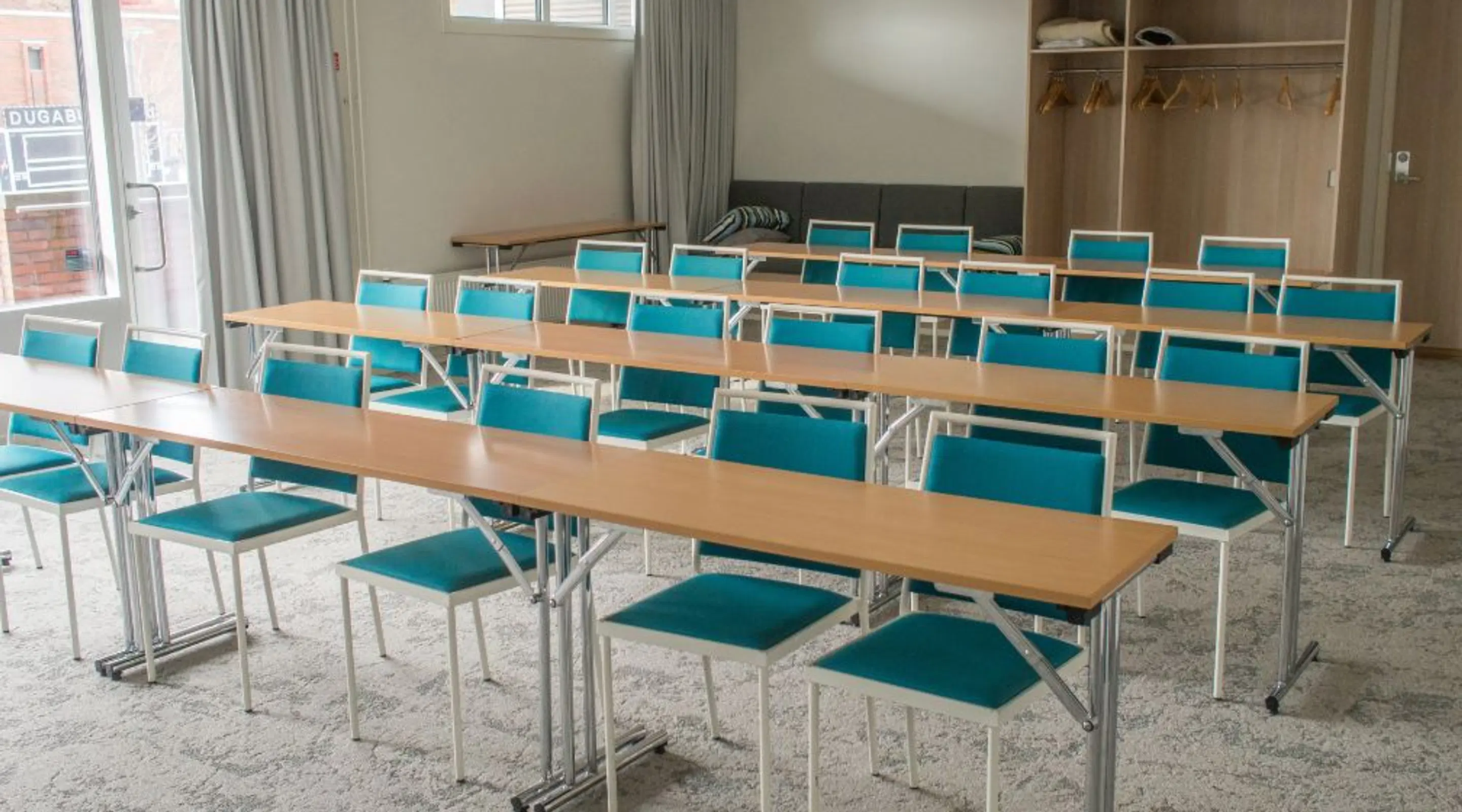 Conference room with long wooden tables and turquoise chairs. Light marble-patterned floor, windows and an open wardrobe in the background.