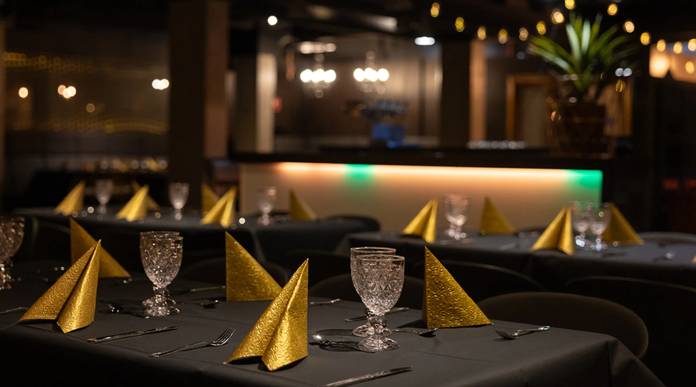 Restaurant tables set with black tablecloths, golden napkins, and crystal goblets. A bar counter with green lighting glows in the background.