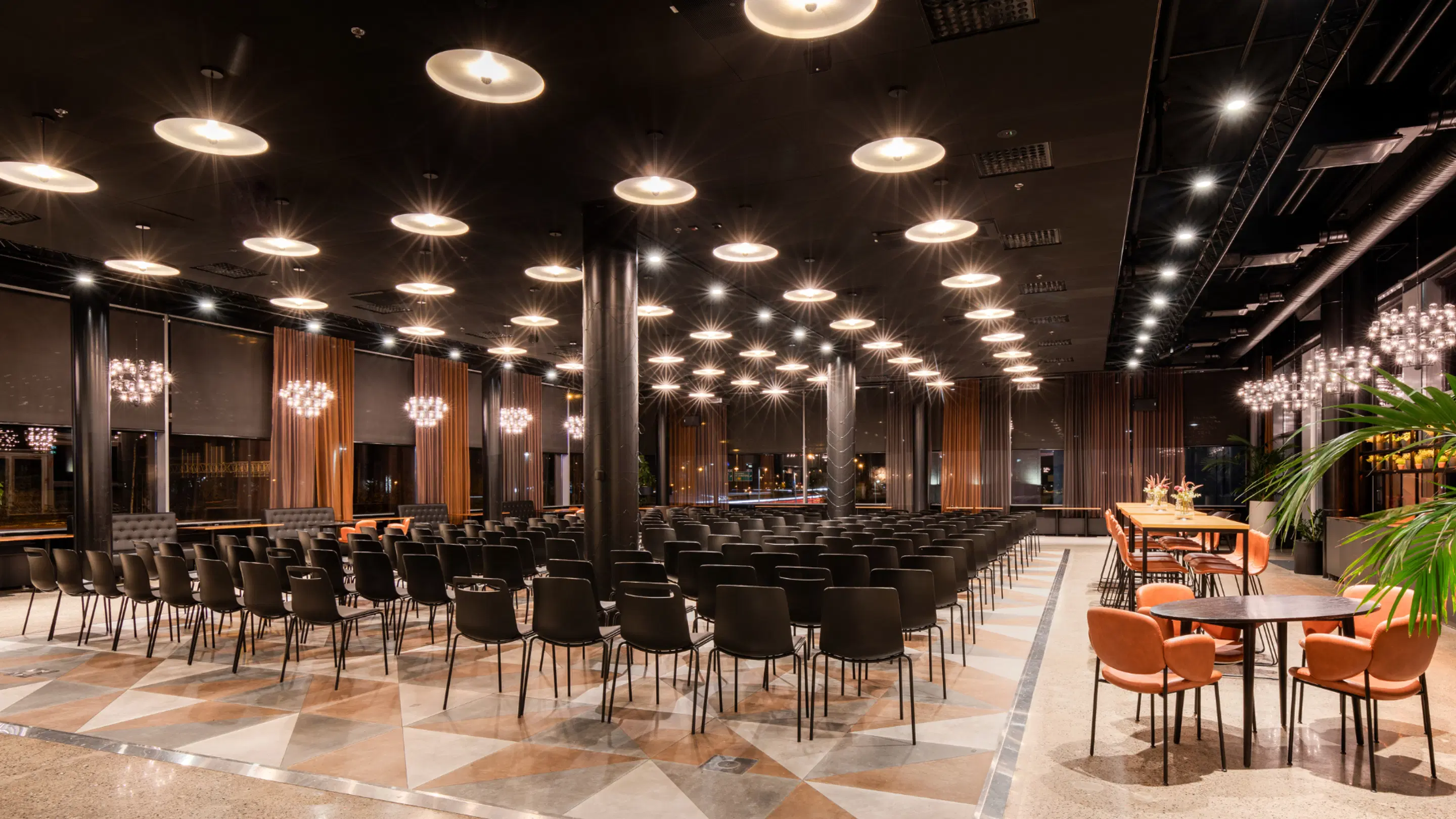 Modern auditorium with rows of black chairs, high ceiling with disc lights, and large windows. Smooth stone floor with orange chairs at tables along the edge.