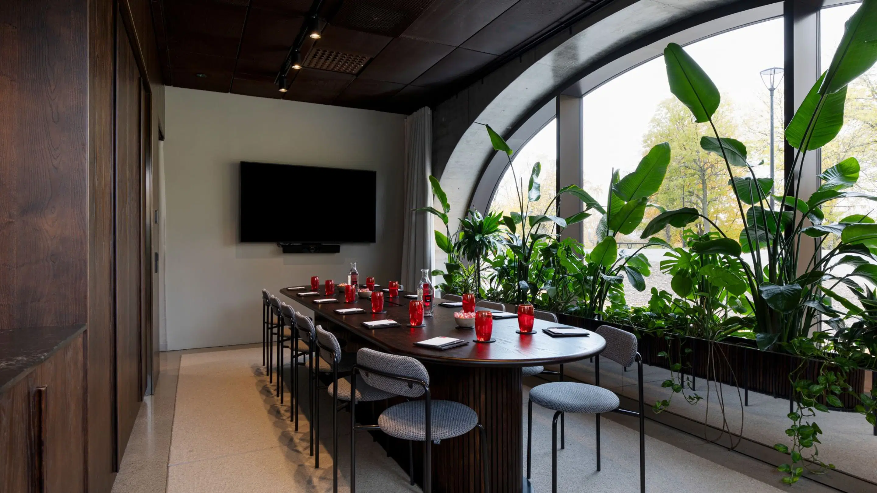Modern meeting room with wooden table, red drinking glasses and abundant greenery in front of arched windows. Television mounted on wall.