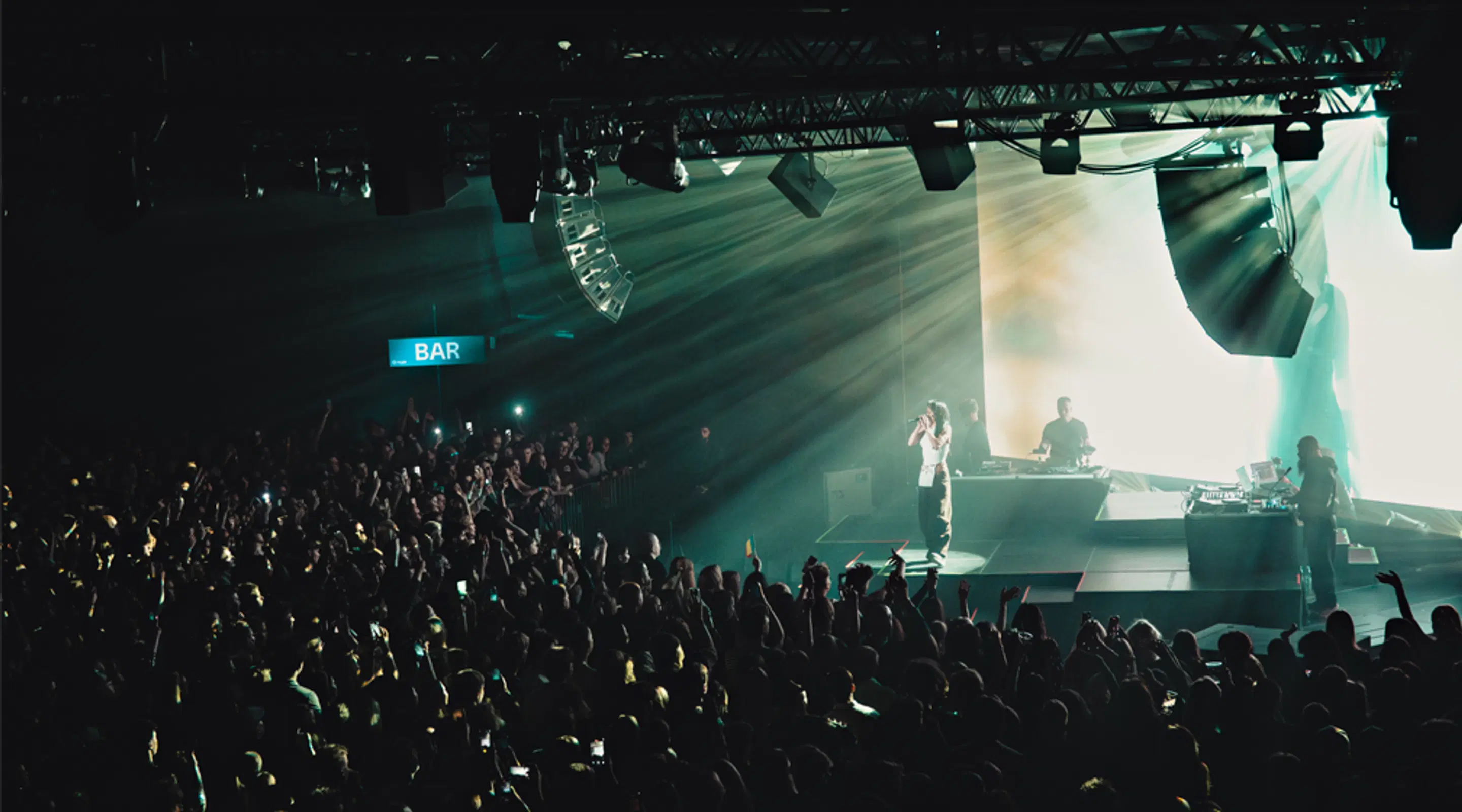 Concert venue with performers on stage and a large audience. Green light beams cut through the dim space, with a bar visible in the background.