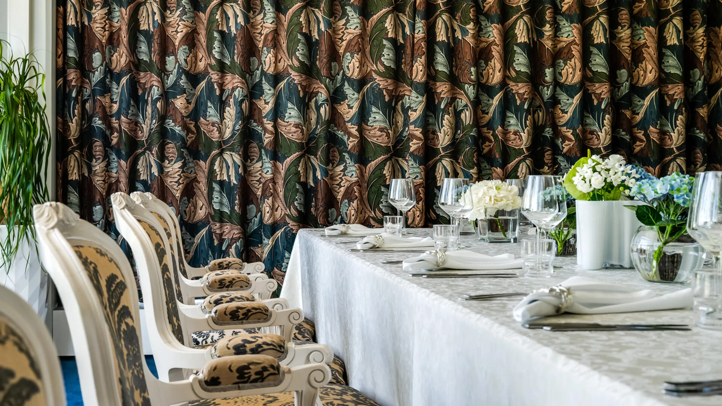 Elegant restaurant table with white tablecloth and flower arrangements. Background features walls covered in leafy patterned fabric and white ornate chairs.