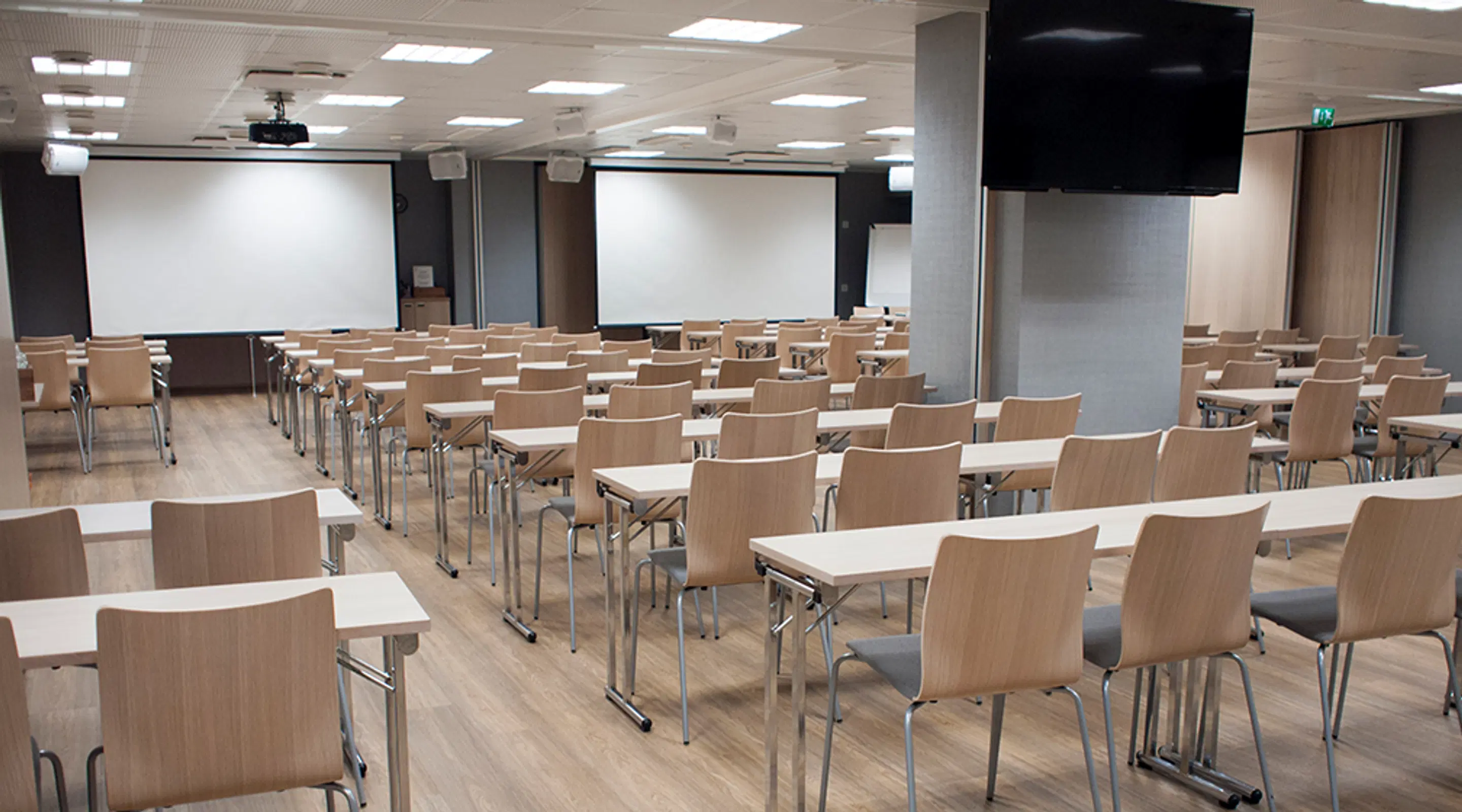 Modernt auditorium med träbord och stolar, två vita dukar och stora skärmar på väggarna, trägolv och bra belysning.