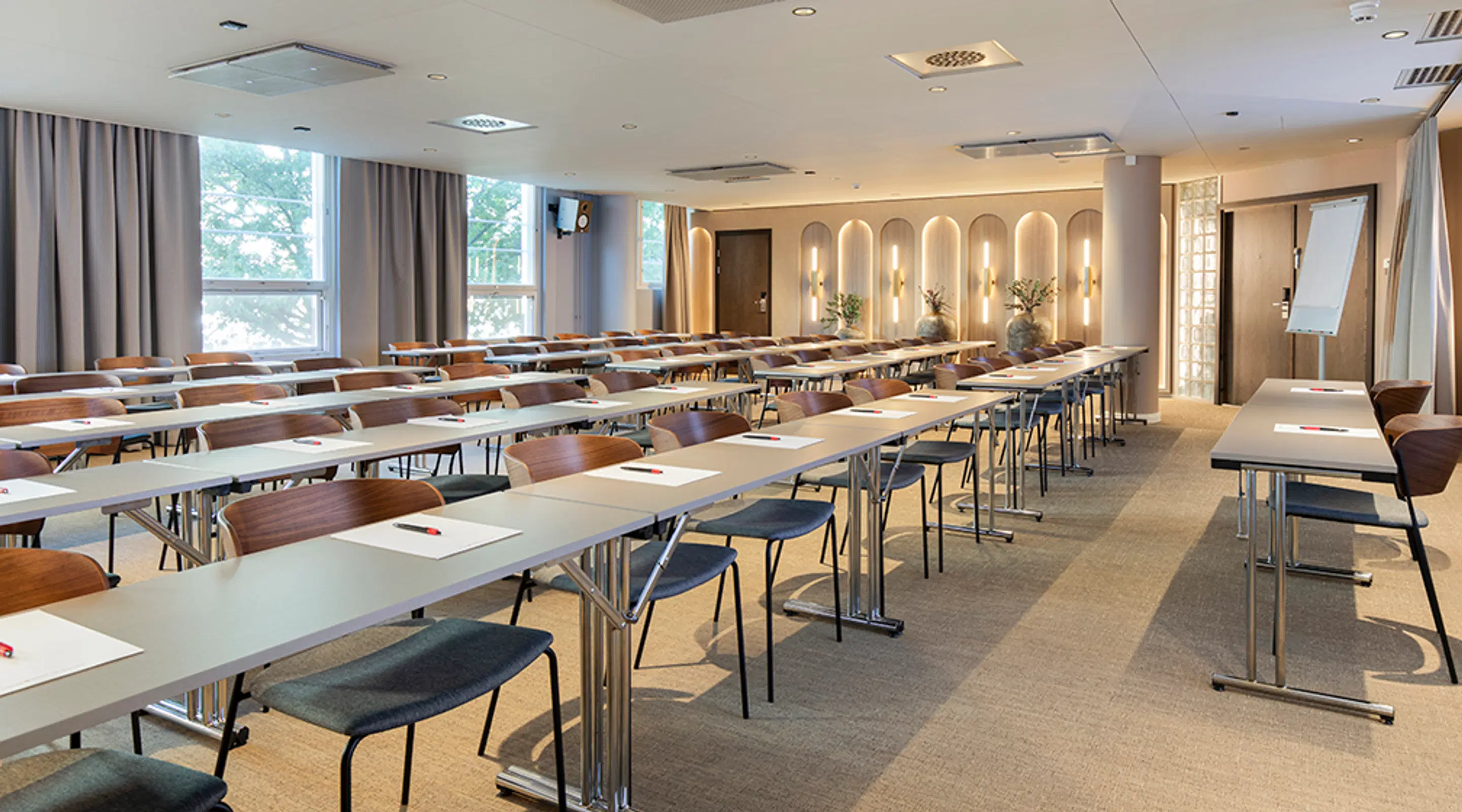 Ljust mötesrum med långa bord i klassrumsformation, dekorativa belysta bågar i bakgrunden och stora fönster.