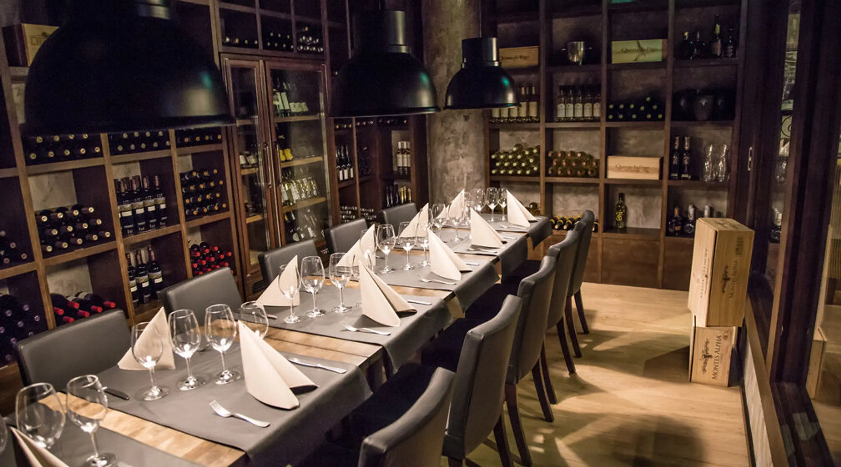 Long table set with wine glasses in restaurant wine cellar. Shelves lined with wine bottles along walls, black pendant lights hanging above.