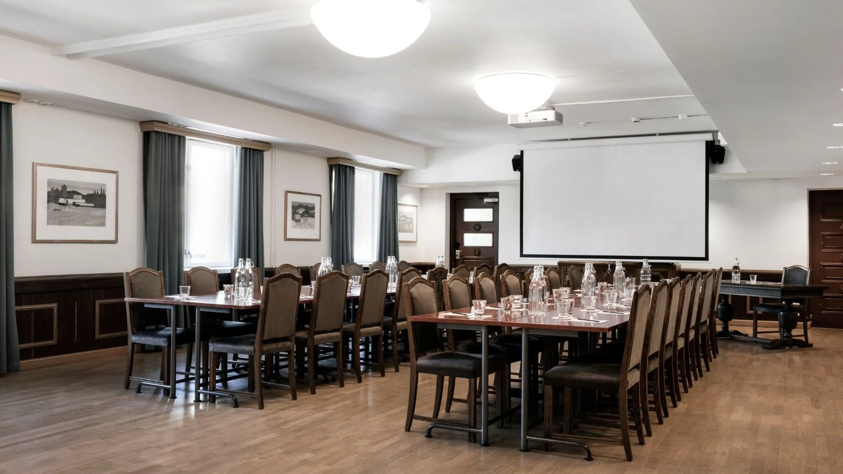 Conference room with tables and chairs, a projector with screen and wooden flooring. The room has large windows with dark curtains.