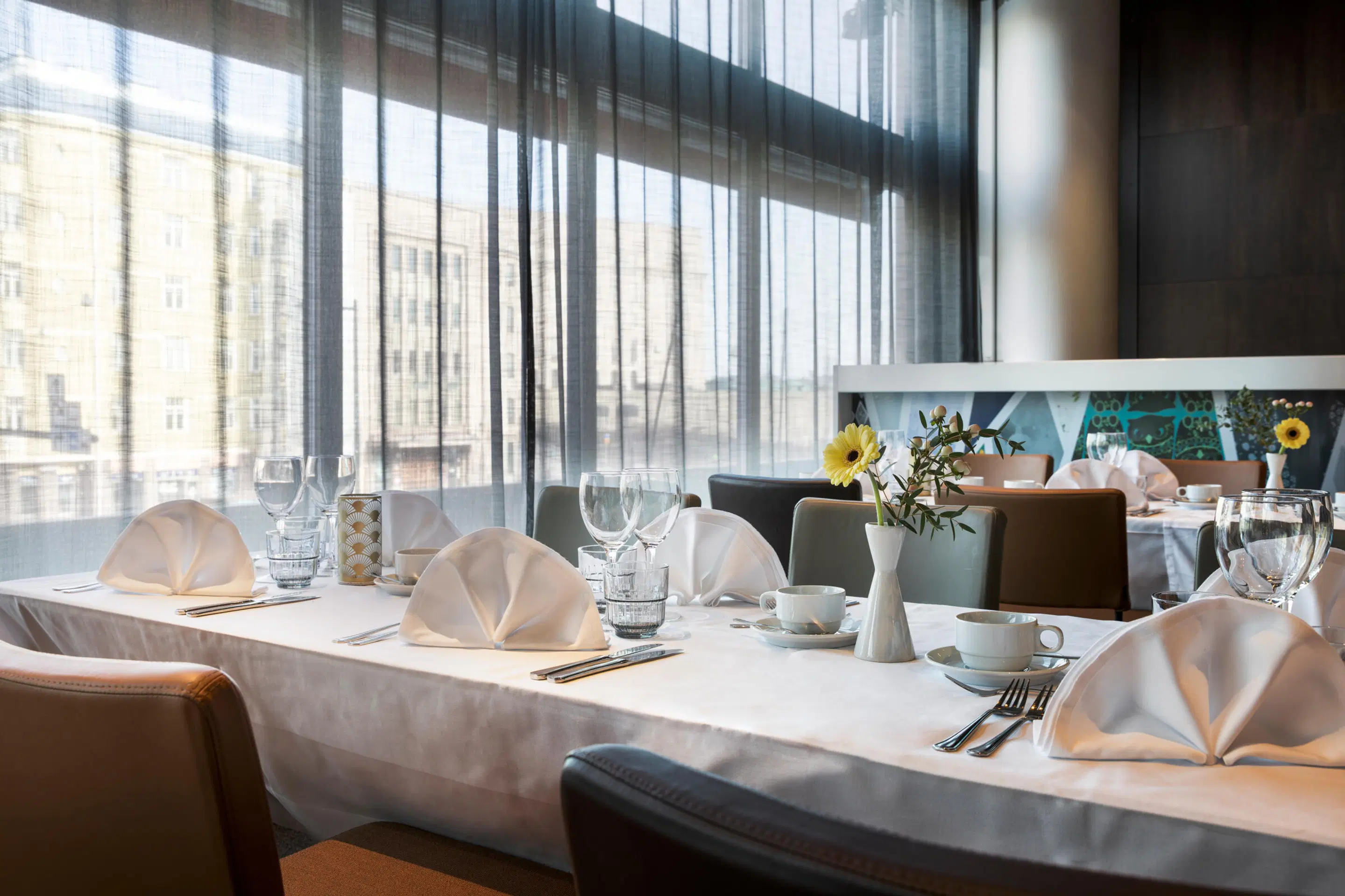Restaurangbord med vit duk, vikta servetter, glas och koppar. Genom stora fönster syns stadsvy, små gula blommor i vas på bordet.