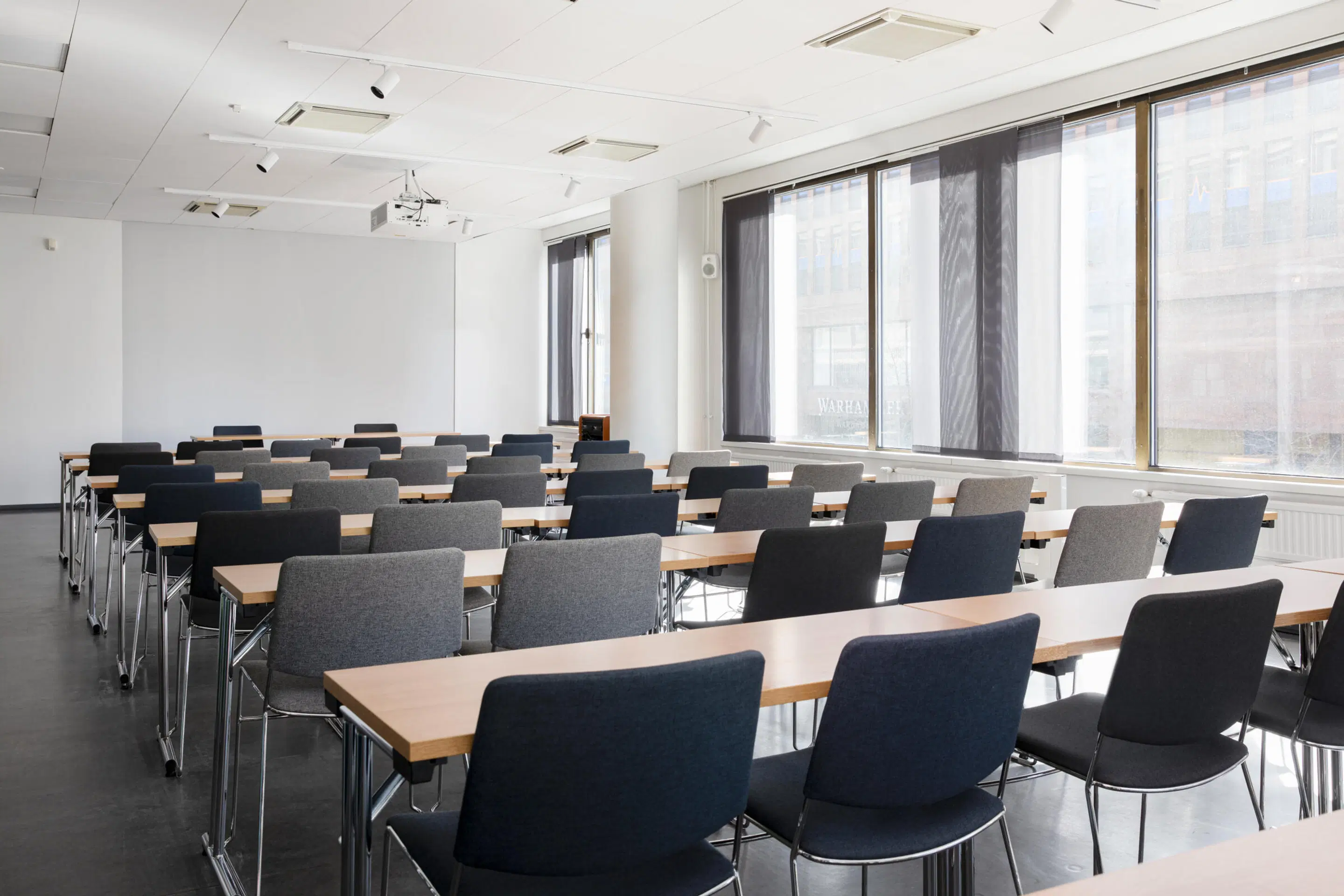 Bright meeting room with light wooden tables and grey chairs arranged in rows, large windows and ceiling-mounted projector. Dark floor.