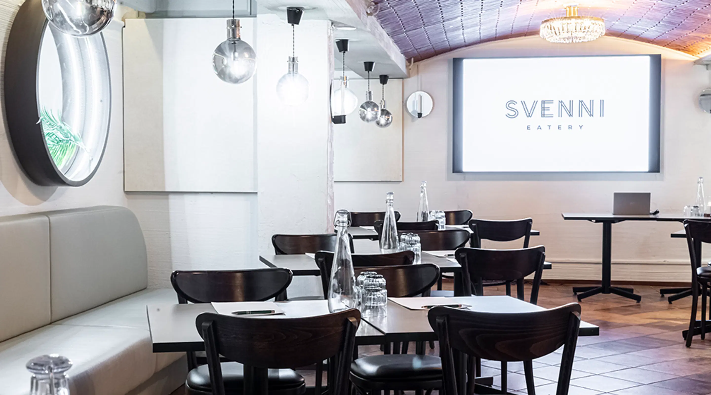 Moderni ravintolan sisätila valkeilla seinillä, mustilla tuoleilla ja tummalla laattalattialla. Kattokruunu ja erilaiset valaisimet luovat tunnelmaa.