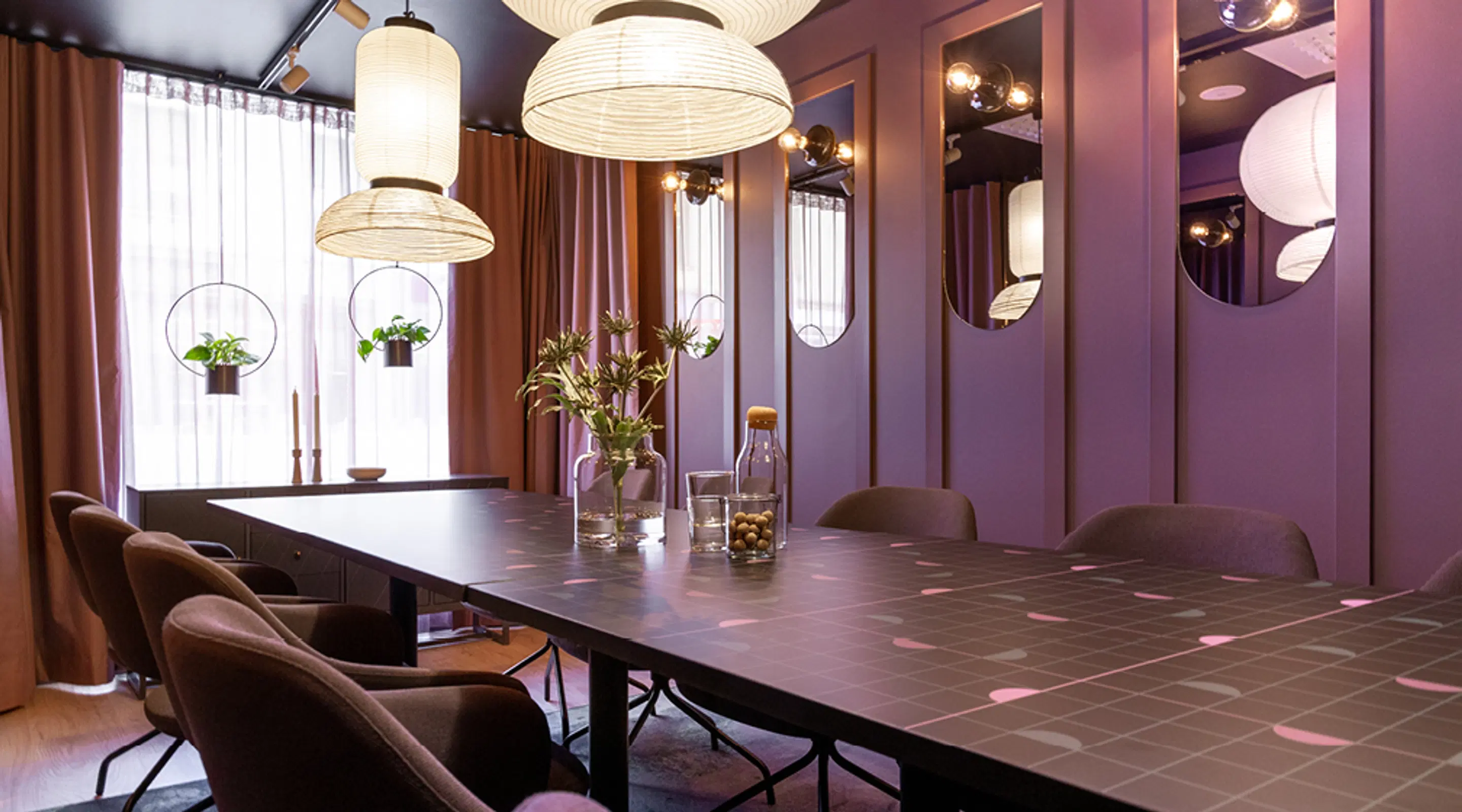 Modern meeting room with purple walls, dark tiled table, comfortable chairs, hanging pendant lights and circular mirrors.