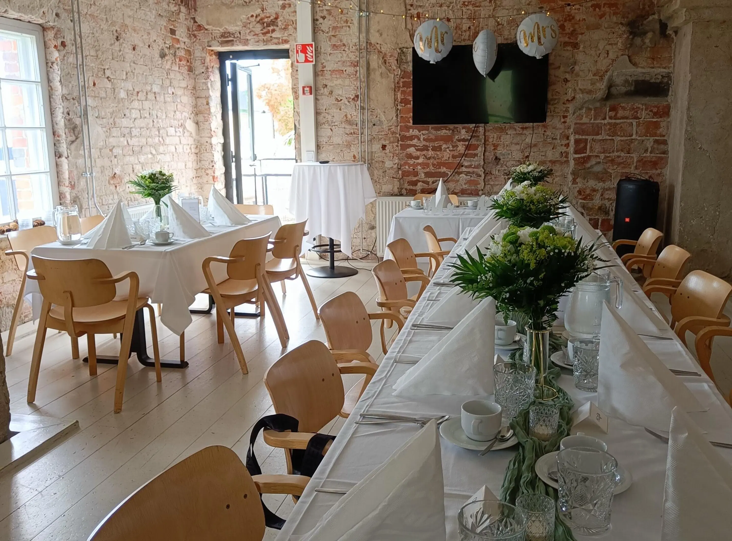 Festlokal med tegelväggar och trägolv. Långa bord med vita dukar och blomsterdekorationer. Trästolar, plant golv och bred ingång.