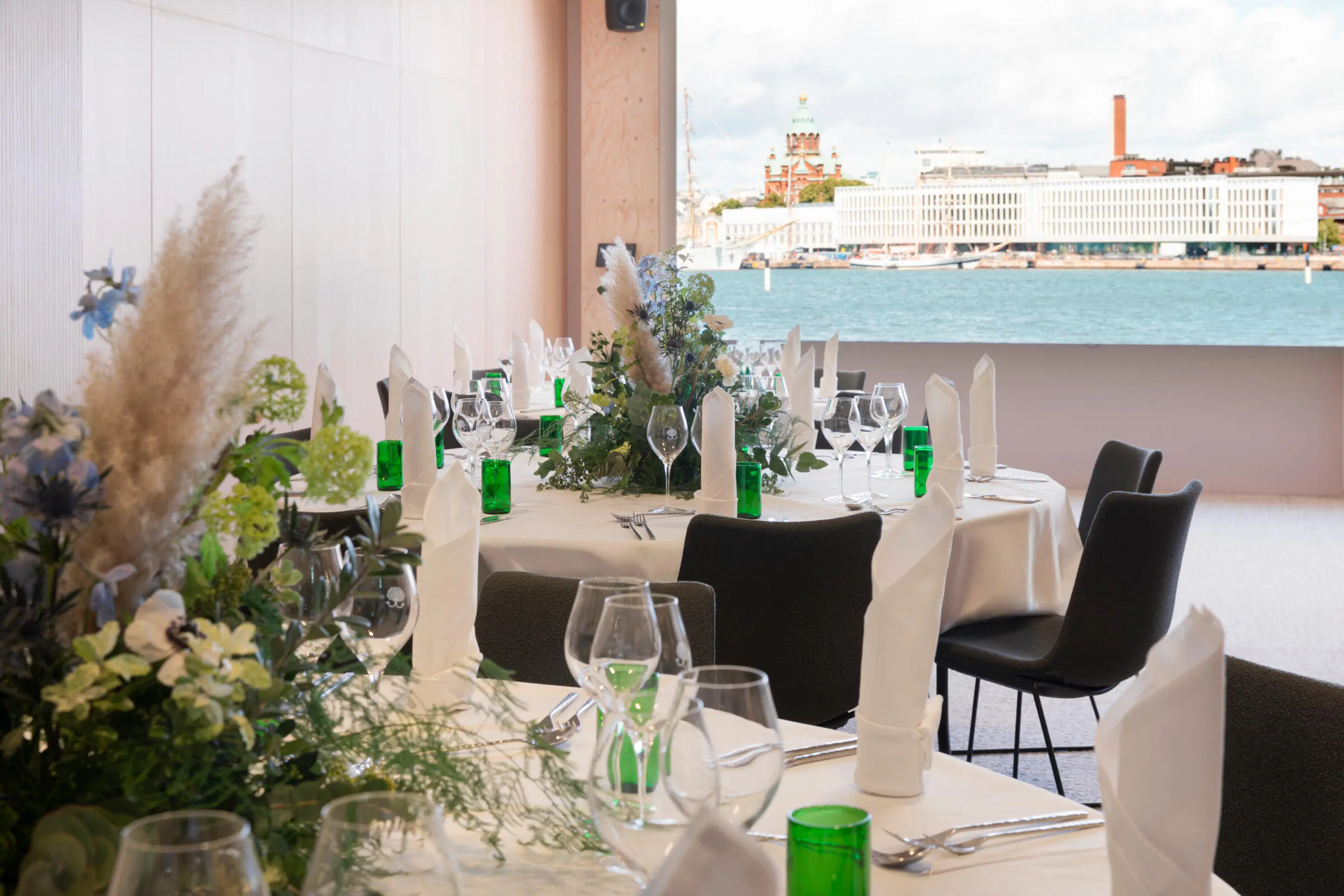Elegantti ravintola merinäköalalla. Pöydät on katettu valkoisilla liinoilla, vihreillä lasipulloilla ja kukka-asetelmilla. Ikkunasta näkyy Helsingin Uspenski-katedraali.