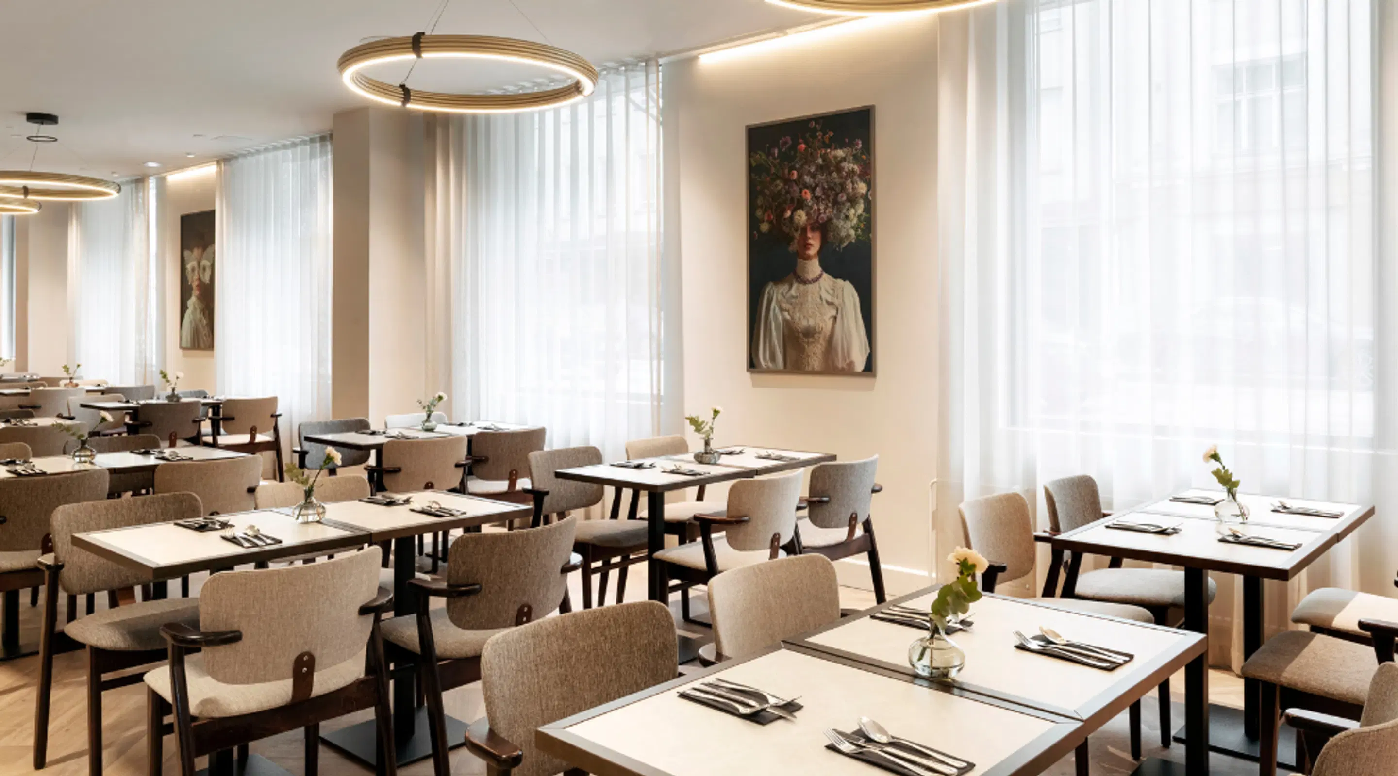 Restaurant dining room with light-colored tables and chairs, large windows, and artwork on walls. Ring-shaped lighting fixtures hang from the ceiling.