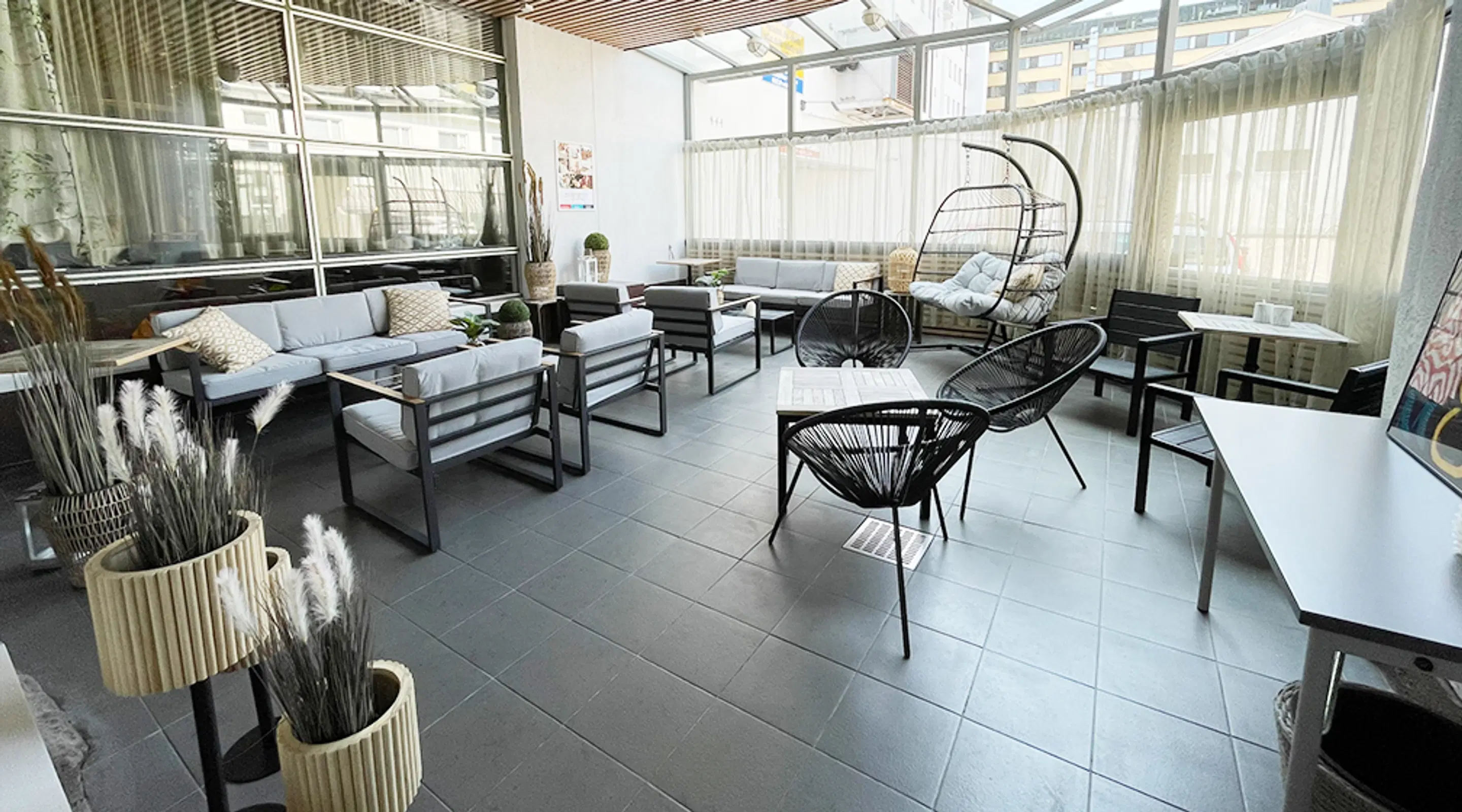 Modern glass-enclosed terrace with stylish furniture: light-colored sofas, black rattan chairs, and a hanging chair. Floor features dark tiles.