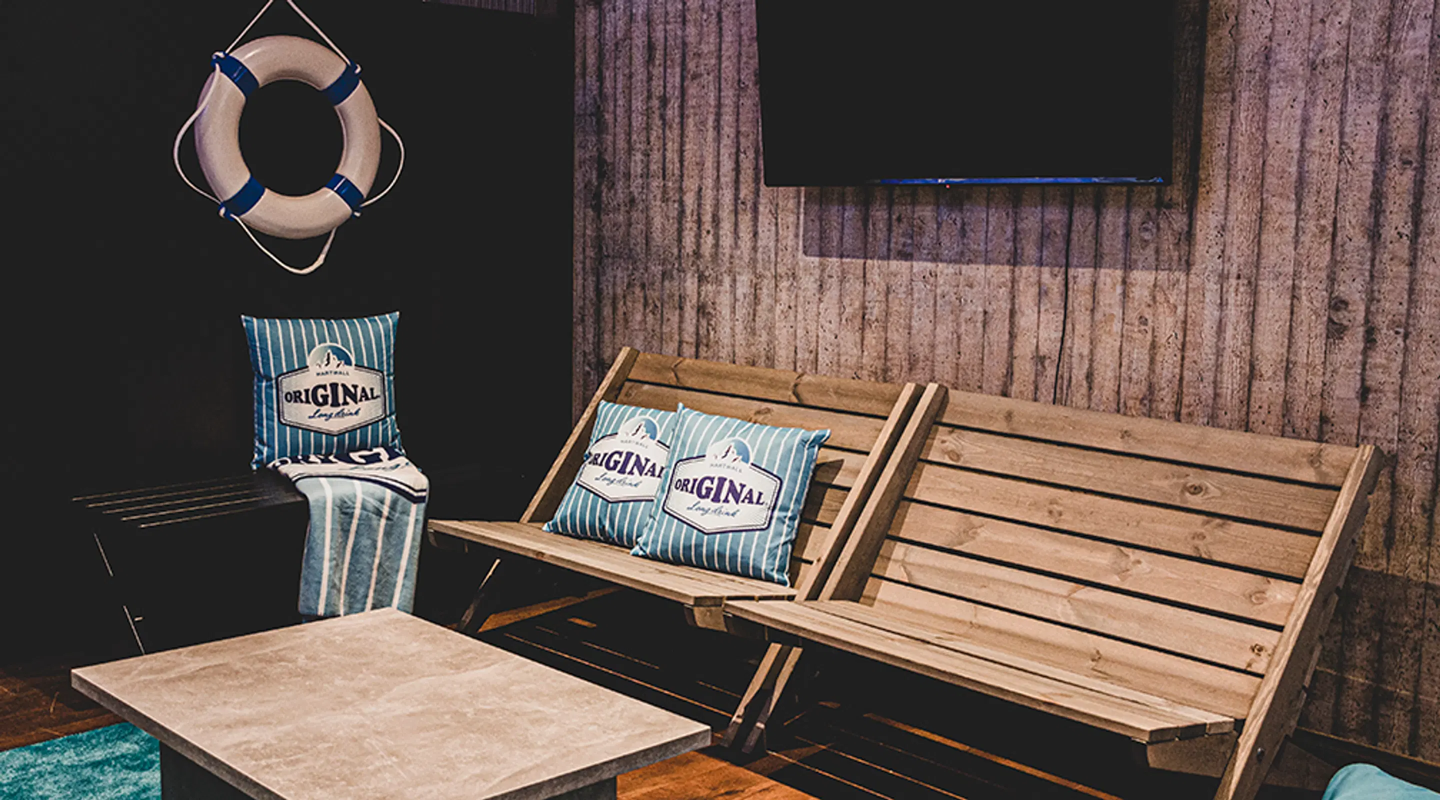 Nautical-themed sauna area with wooden walls. Wooden bench, blue-striped pillows and a life preserver hanging on the wall.