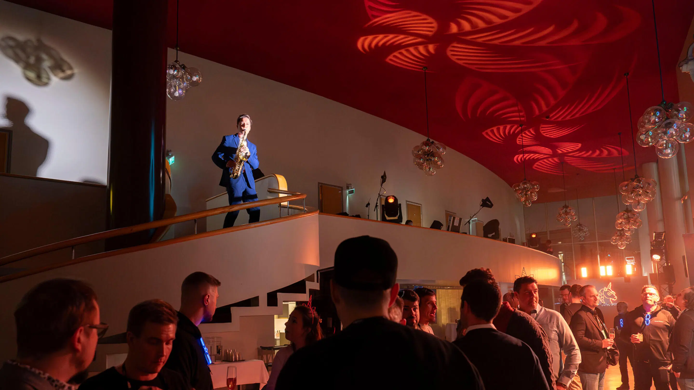Saxophonist in blue suit performing on upper balcony while audience watches from below. Red lighting patterns on ceiling and hanging crystal chandeliers.