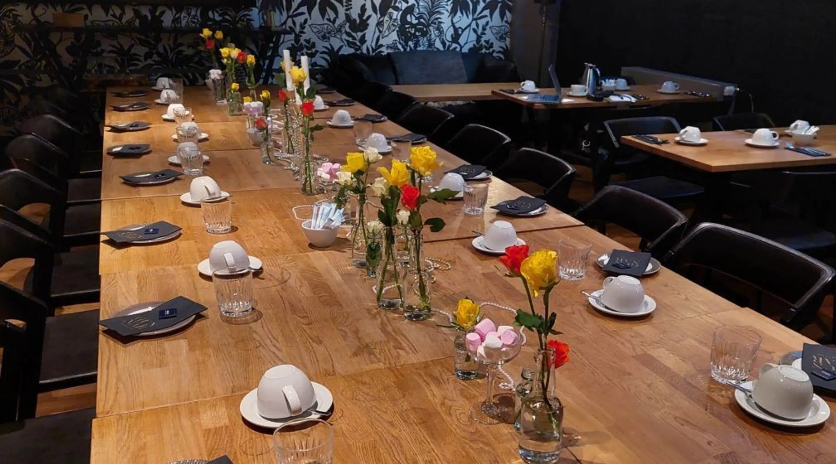 Meeting room with long wooden table decorated with flower arrangements, white coffee cups and glasses. Dark floral wallpaper and additional tables in background.