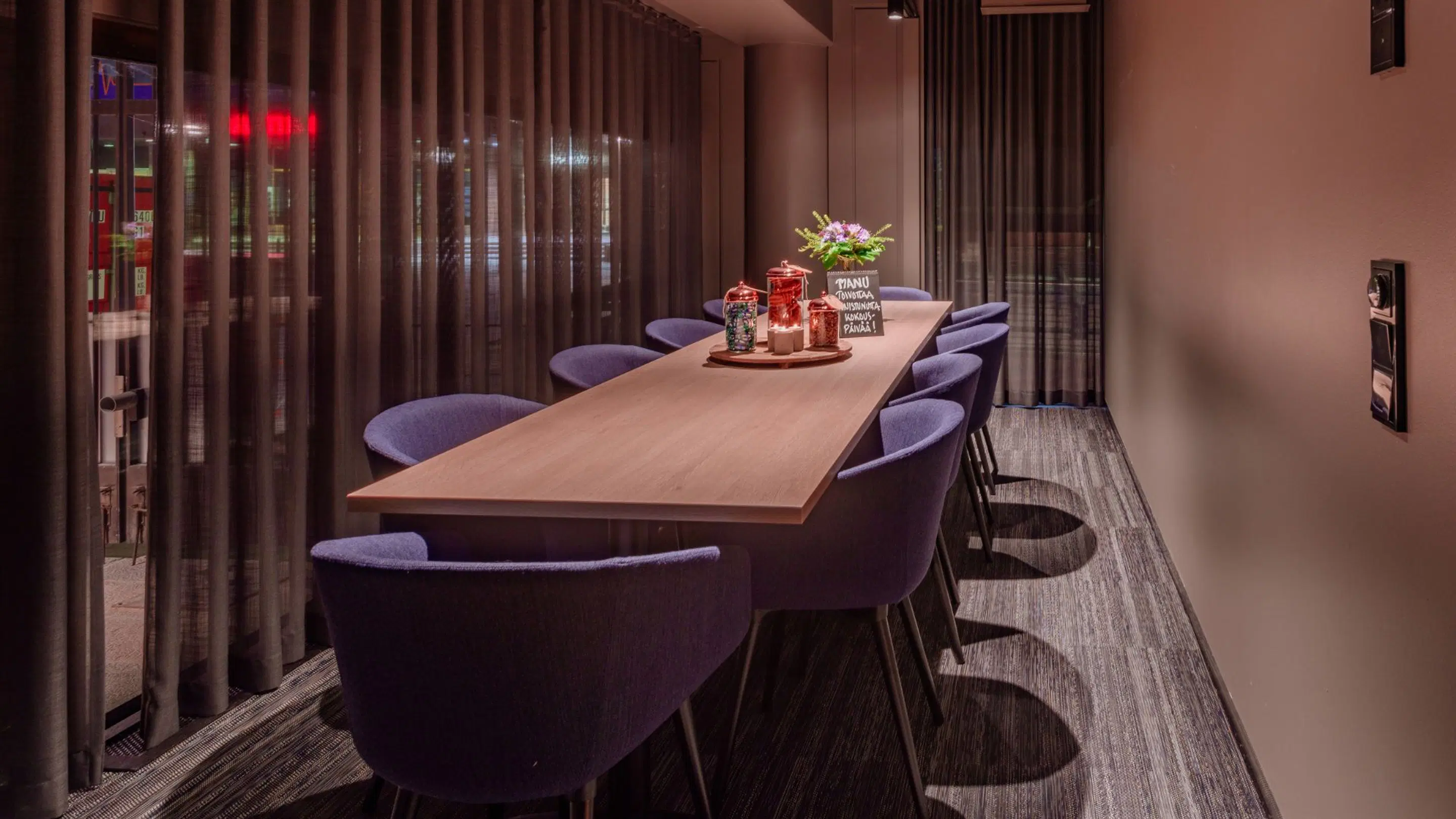 Meeting room with a long wooden table, purple chairs and a flower arrangement with decorations in the center. The room has dark curtains and textured flooring.