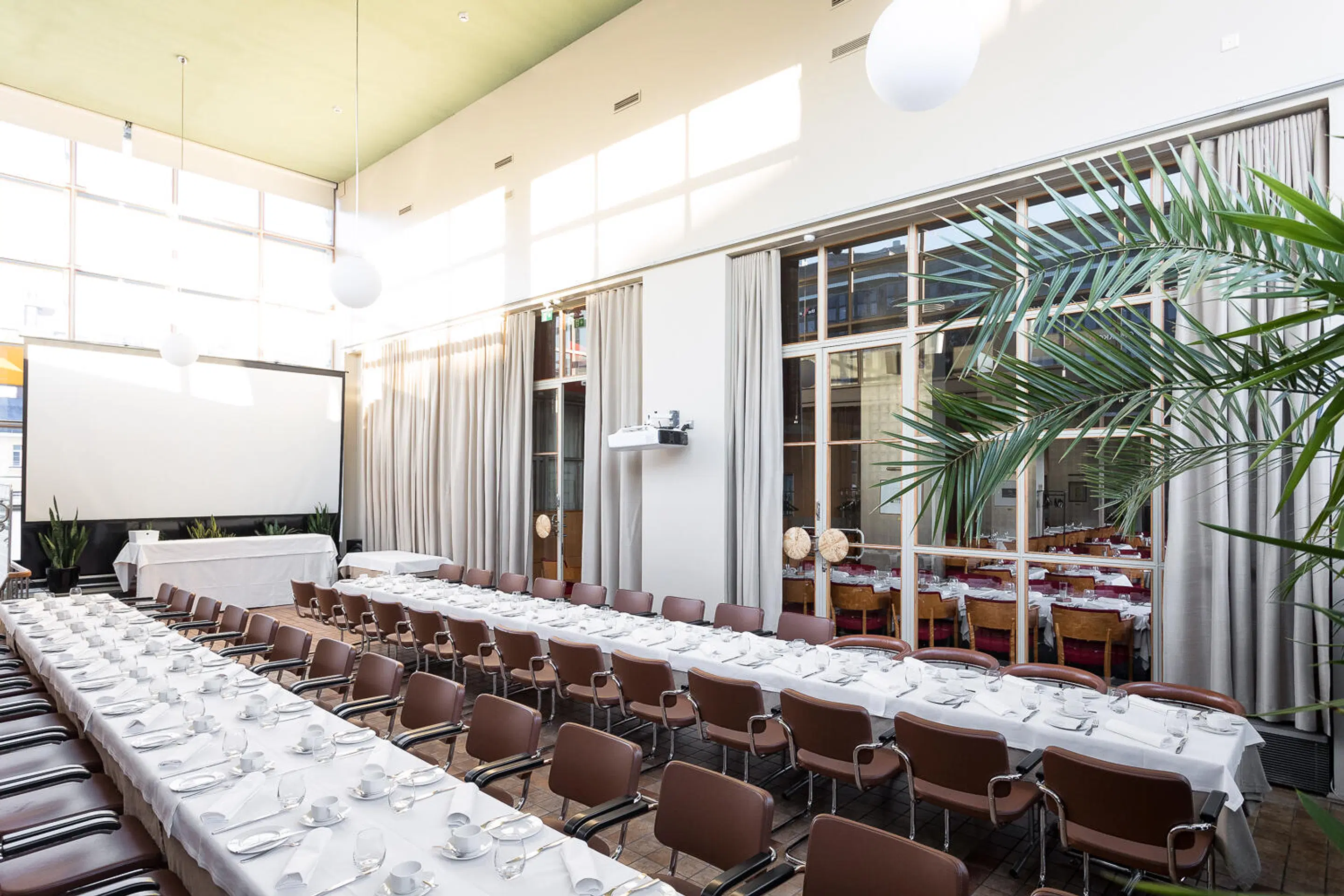 Bright meeting room with long tables covered in white tablecloths and place settings. Brown chairs, projection screen and large windows. Green palm plant in corner.