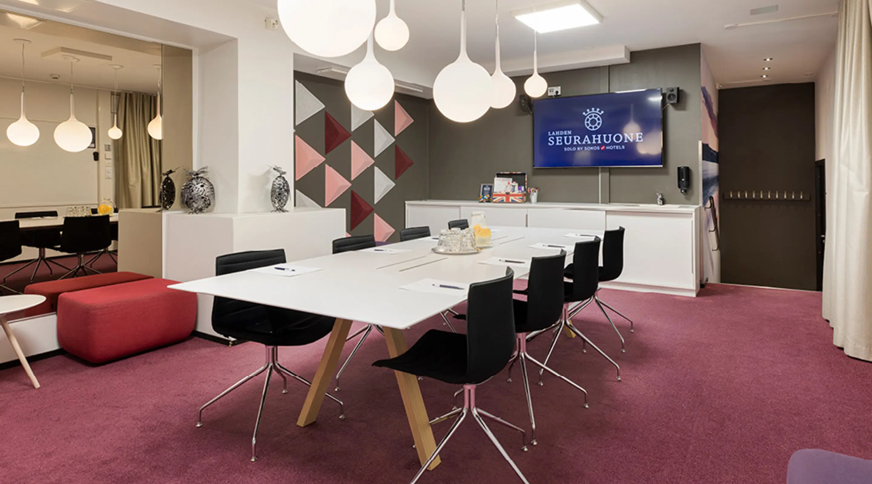 Modernt konferensrum med vitt bord, svarta stolar, rosa heltäckningsmatta och hängande armaturer.