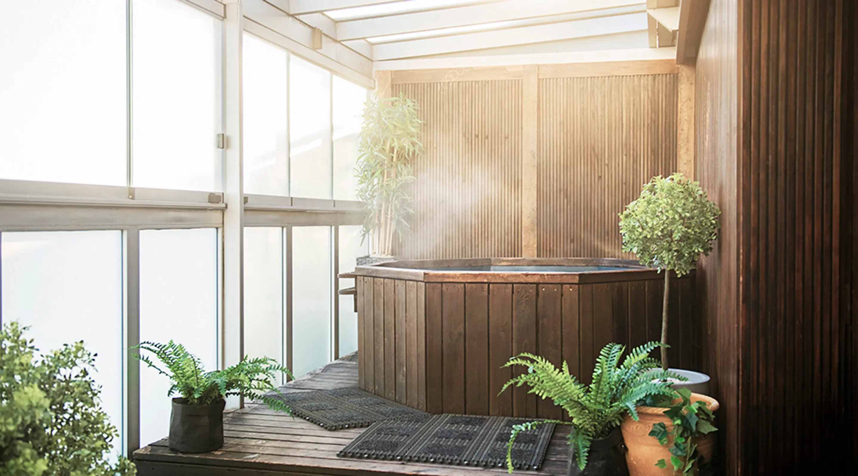 Wooden hot tub indoors surrounded by potted greenery and ferns. Bright space with glass wall and wooden flooring.