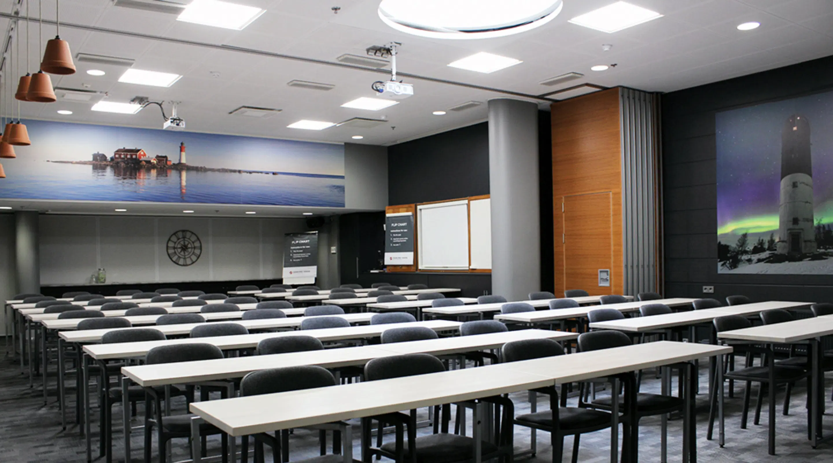 Auditorium with long tables and chairs, illuminated by ceiling lights. Walls feature seascape and northern lights artwork.