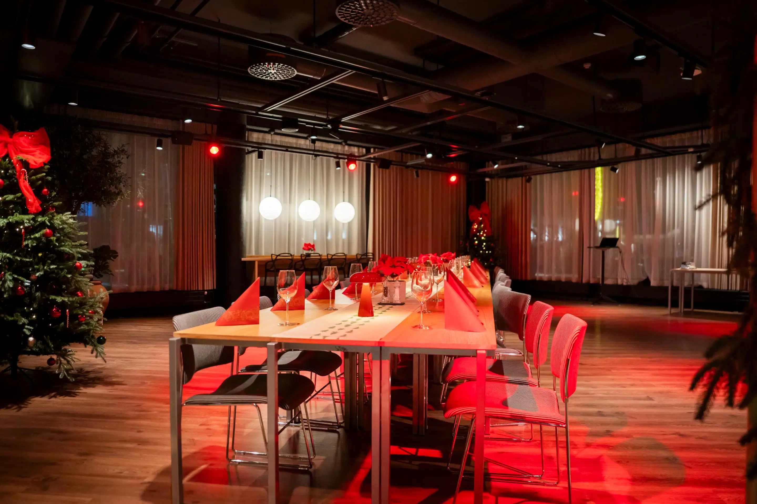 Event space with a table set with red napkins, decorated Christmas tree, and wooden floor. Room features red lighting and white curtains.
