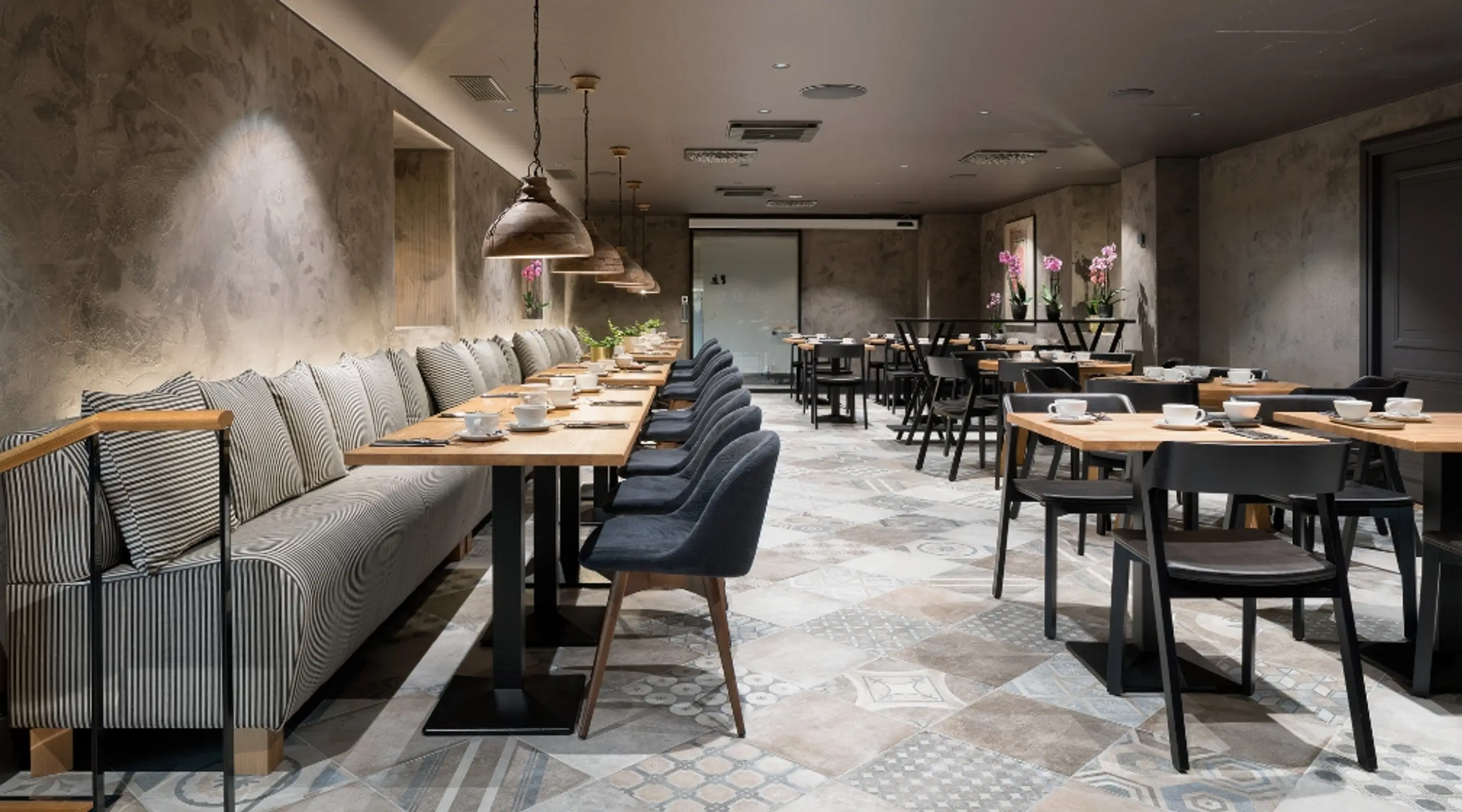 Modern restaurang med betongväggar, lång randig soffa och träbord. Utrymmet har ett mönstrat klinkergolv och industriella hänglampor.