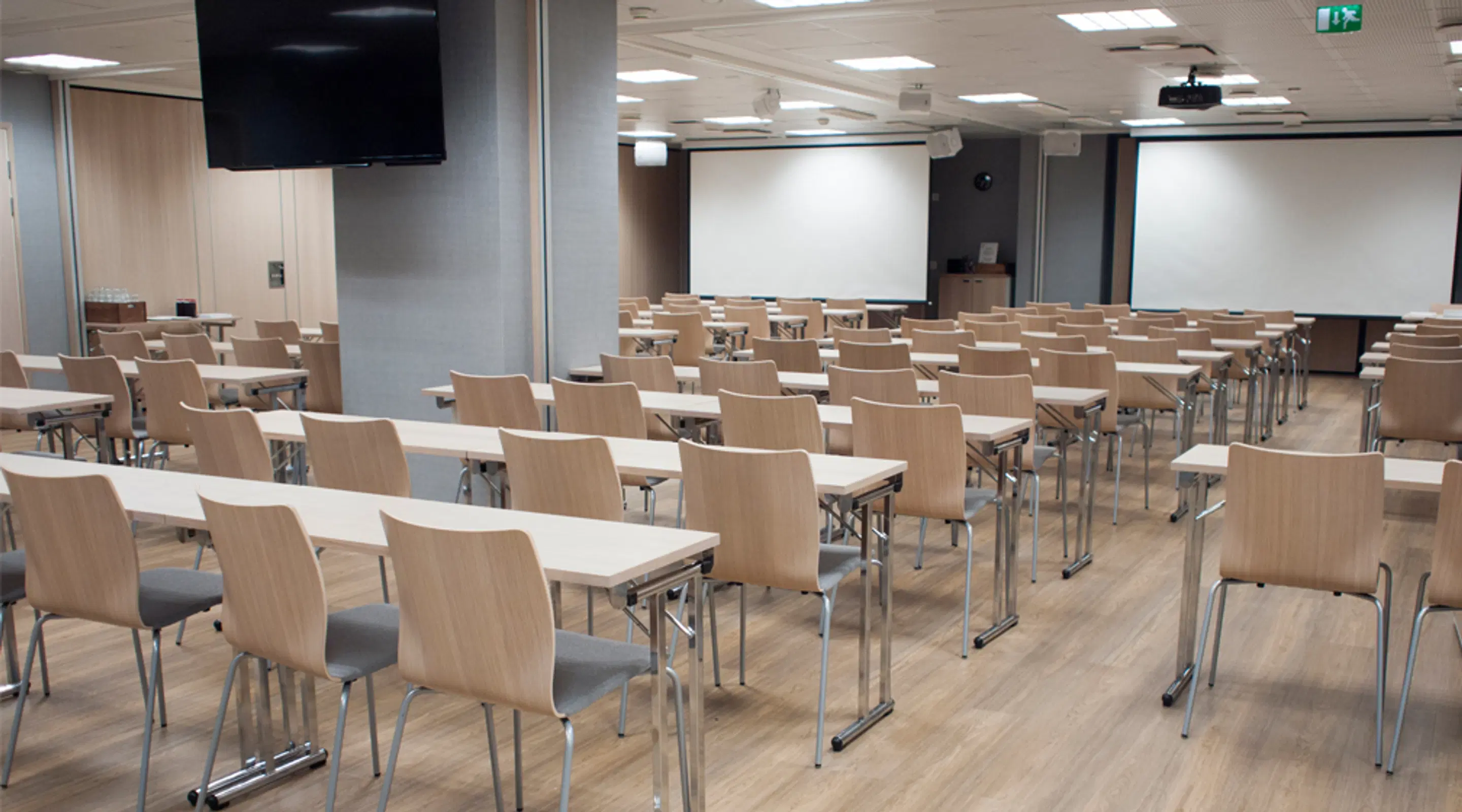 Modern auditorium med ljusa träfärgade stolar och vita bord i rader. Projektorduk framme och TV-skärm i taket. Ljust trägolv och bra belysning.