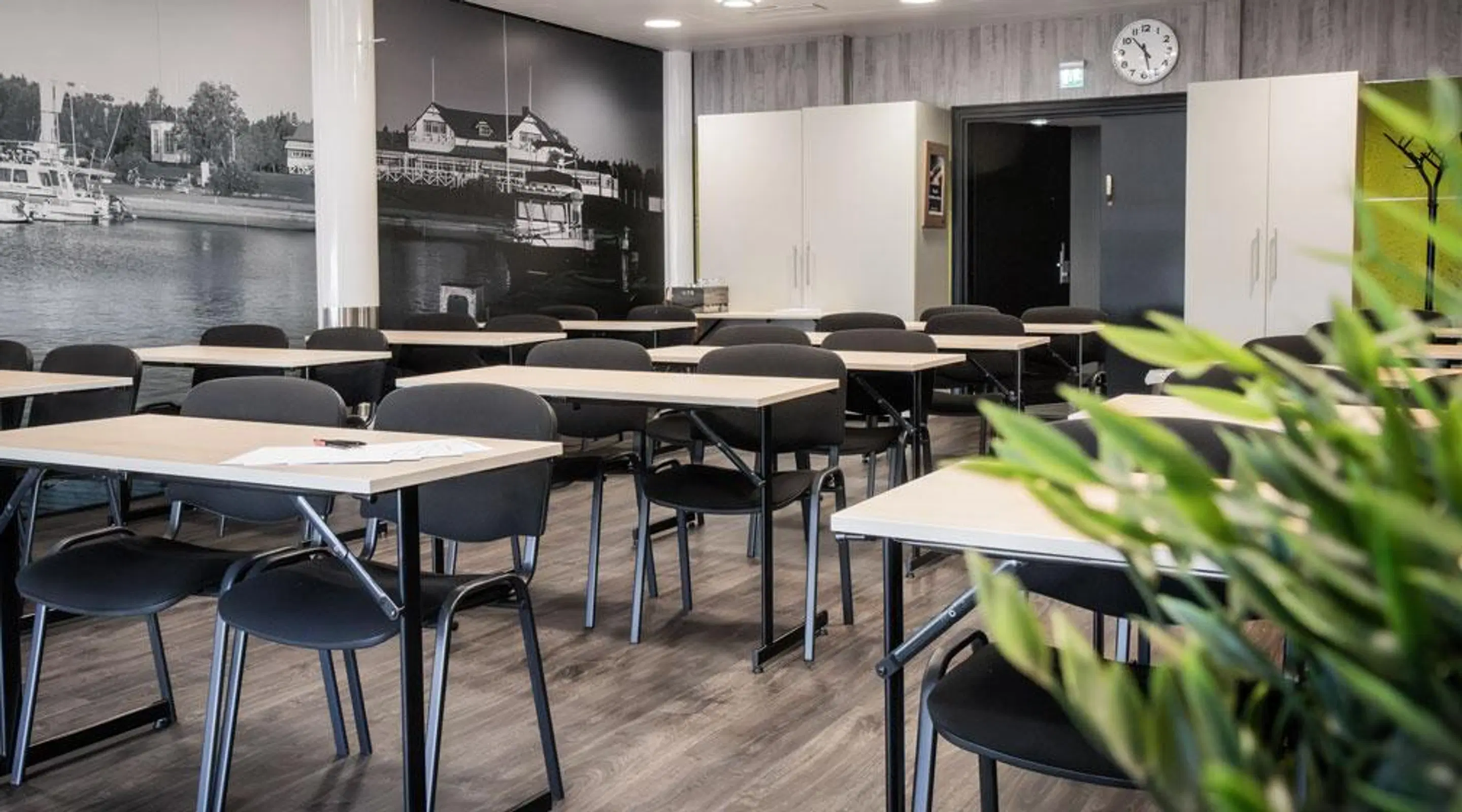 Training room with light-colored tables and black chairs. Background features a harbor photo mural and hard flooring.