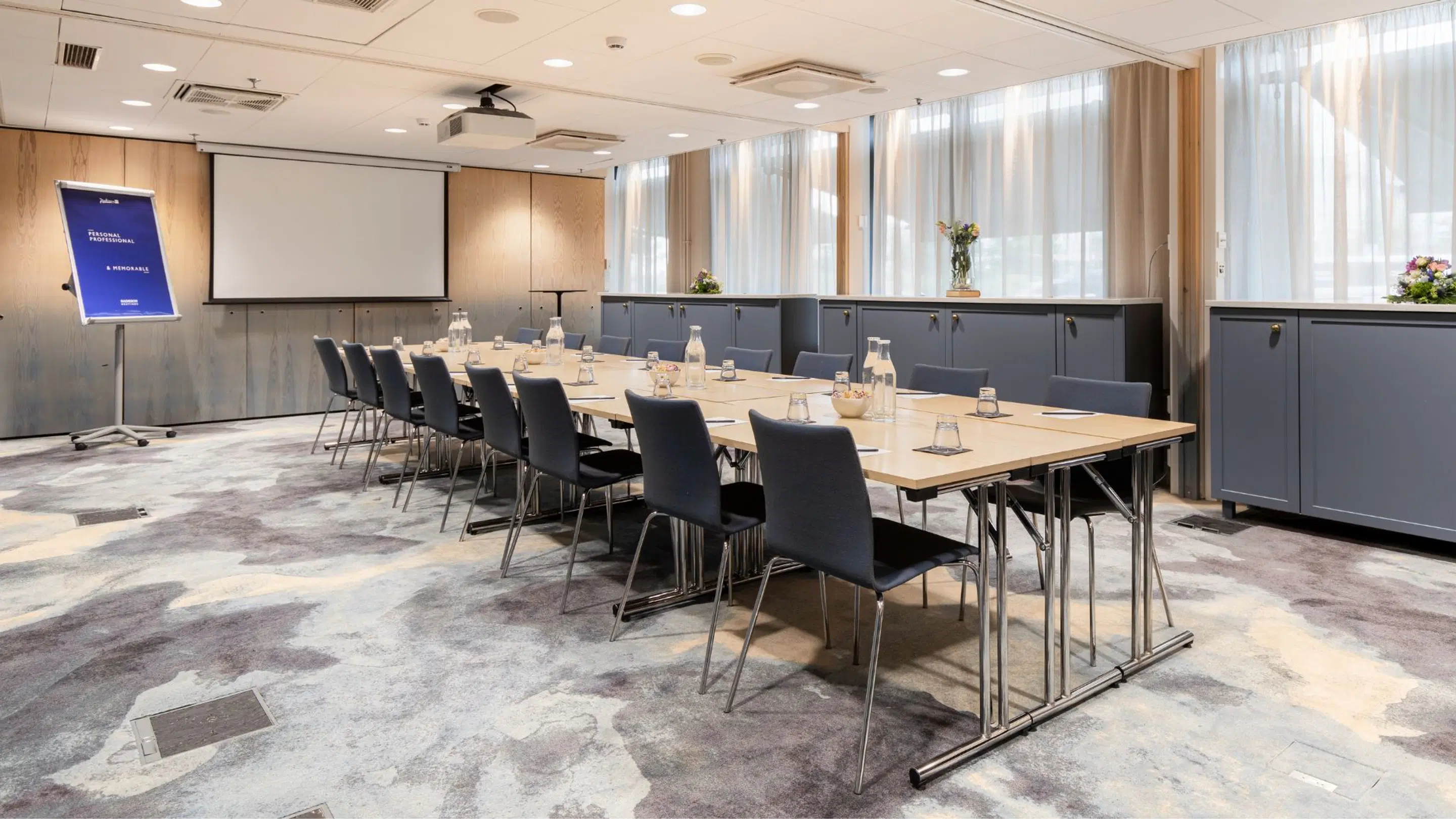 Konferensrum med långt bord och mörkgrå stolar, projektorduk och projektor. Stora fönster, träpaneler på väggarna och marmormönstrat golv.