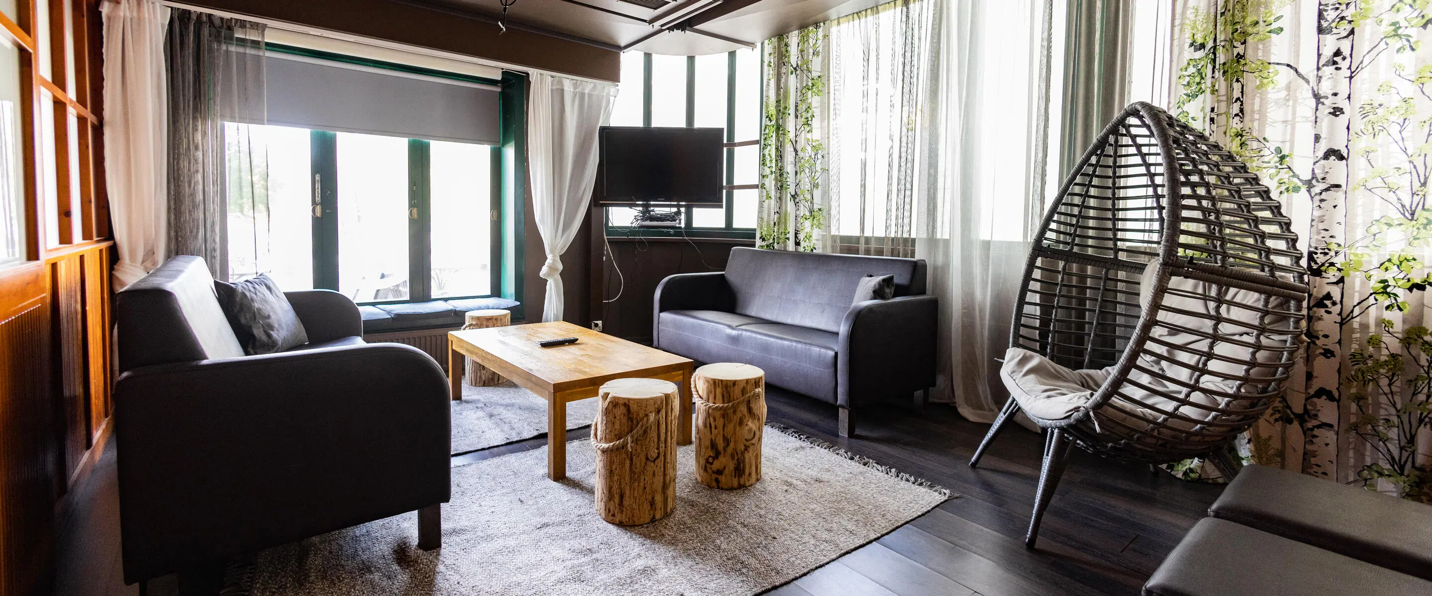 Modern living room with sofas, wooden table, hanging egg chair and large windows. Floor is hard wood with thin rugs.